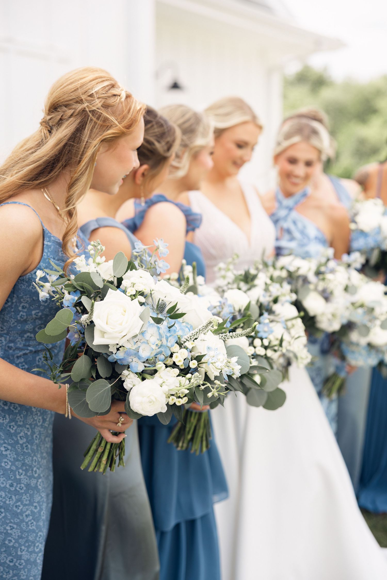Bridesmaids in dusty blue dresses hold white and blue floral bouquets with eucalyptus greenery beside the bride outdoors.