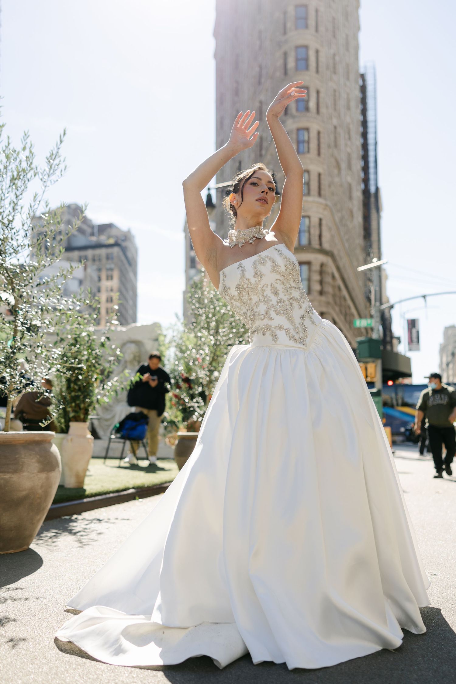 Bride in a satin ball gown with beaded bodice dances with raised arms before the Flatiron Building in New York City.