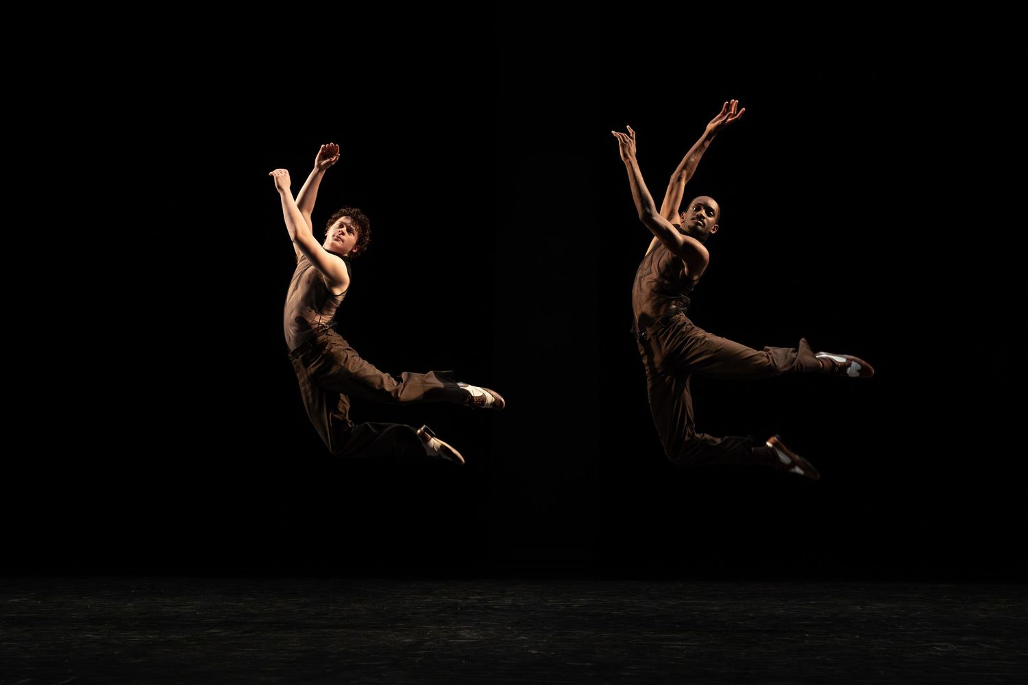 Two dancers leap in mid-air with arms stretched upward against a black background in a powerful stage performance.