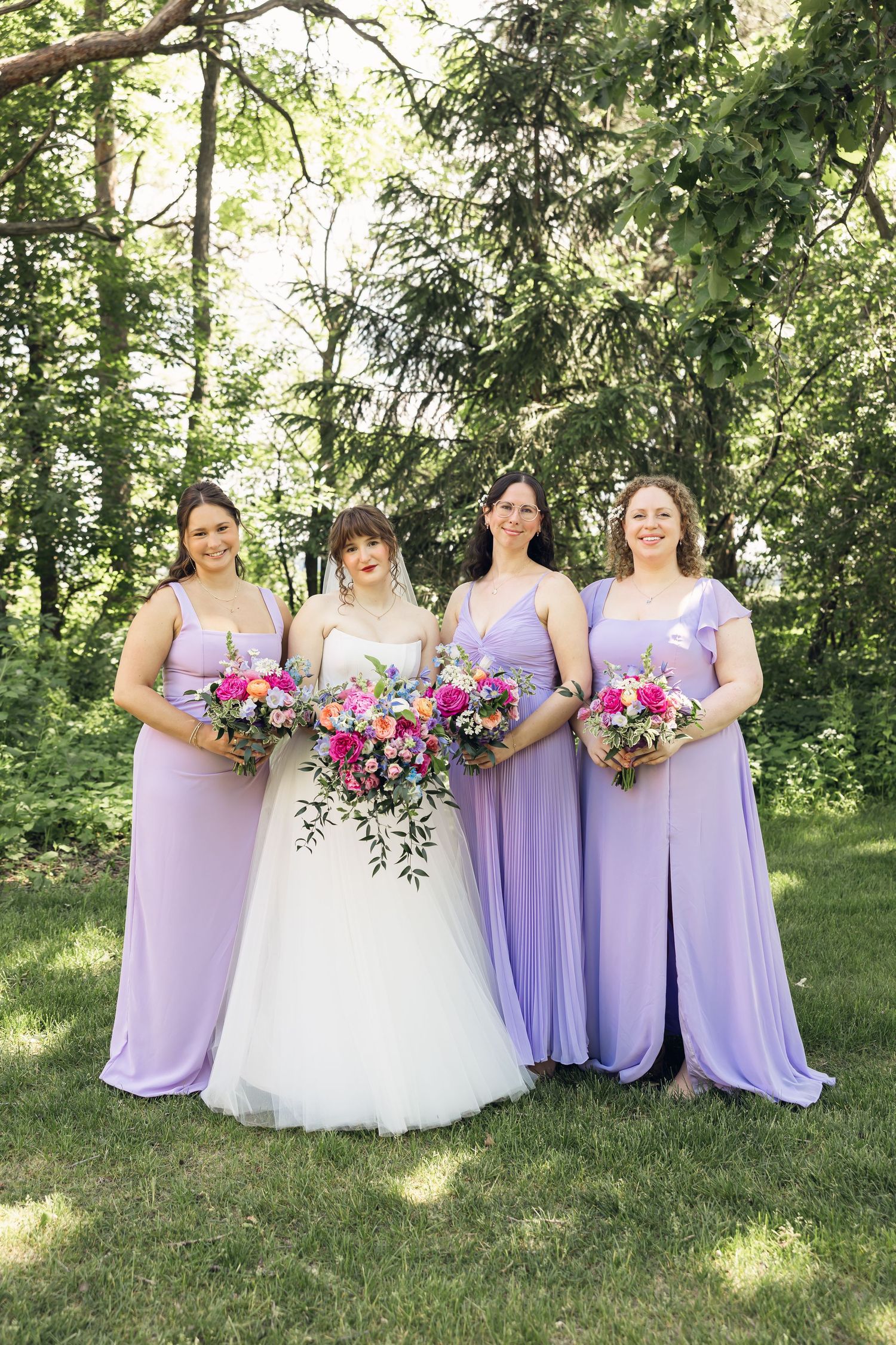 Bride in a white gown stands with three bridesmaids in lavender dresses, holding colorful bouquets in a sunny wooded setting.