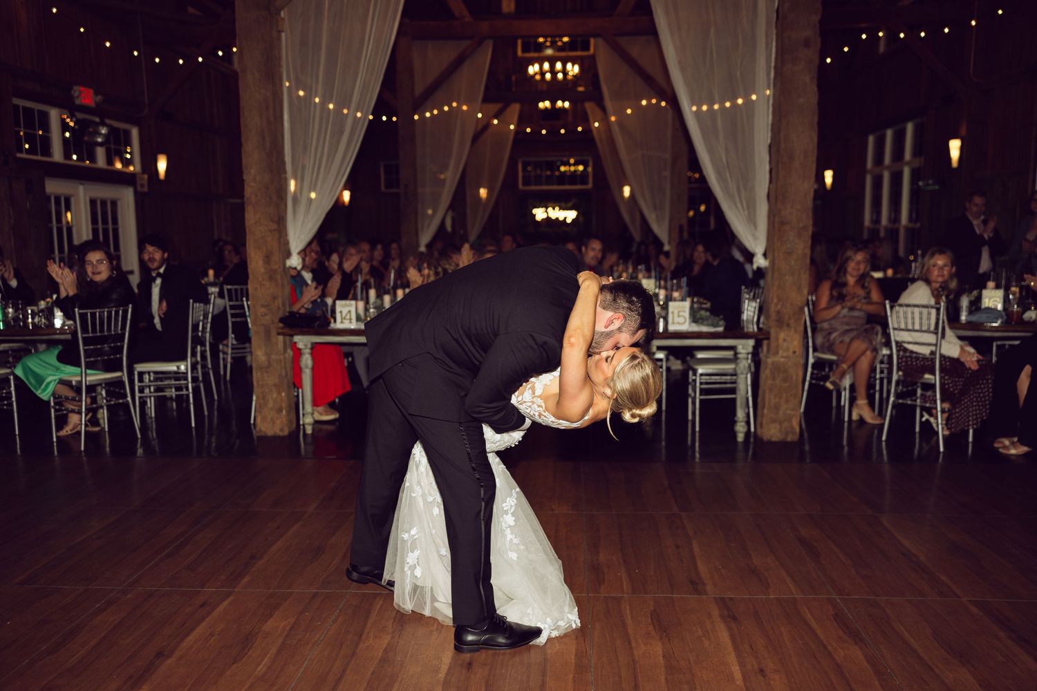 A groom dips his bride during their first dance on a wooden floor in a rustic barn venue with draped fabric and string lights.