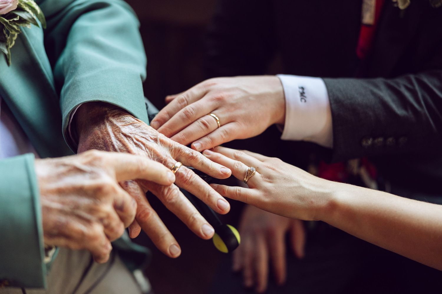 Close-up of an elderly man's hand joined with the newlyweds' hands, showing wedding rings and a green suit sleeve.
