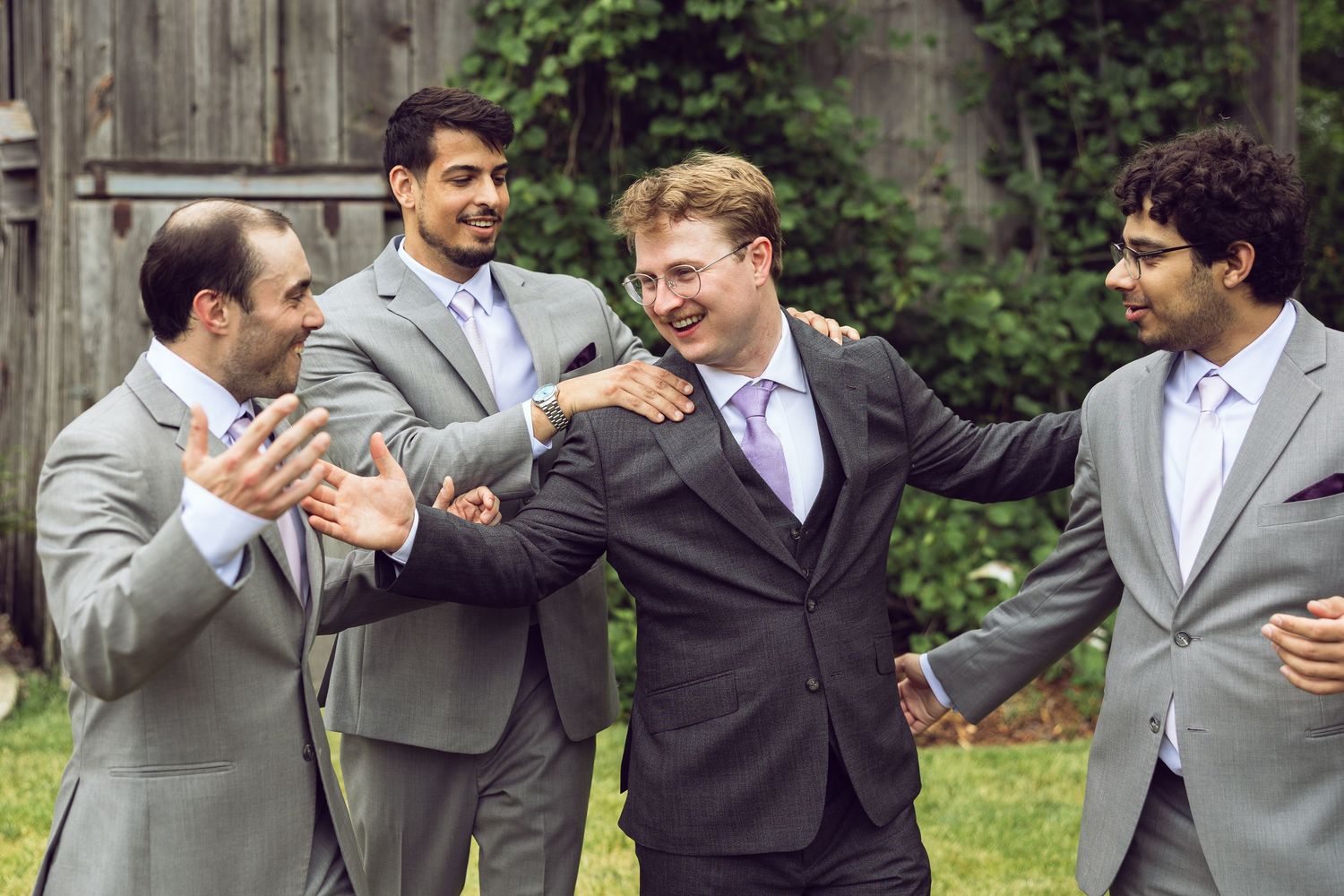Groomsmen in gray suits laugh and joke with the groom in a lavender tie during a lighthearted outdoor wedding moment.