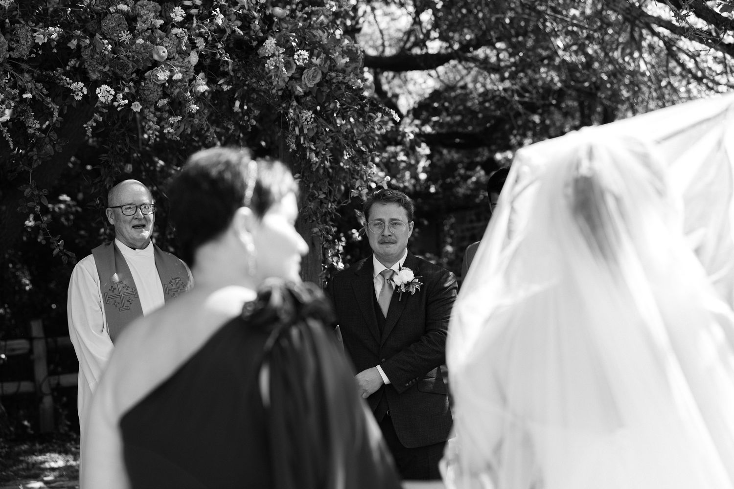 Black and white photo of a smiling groom and officiant at an outdoor wedding ceremony as the bride's veil blows in the foreground.