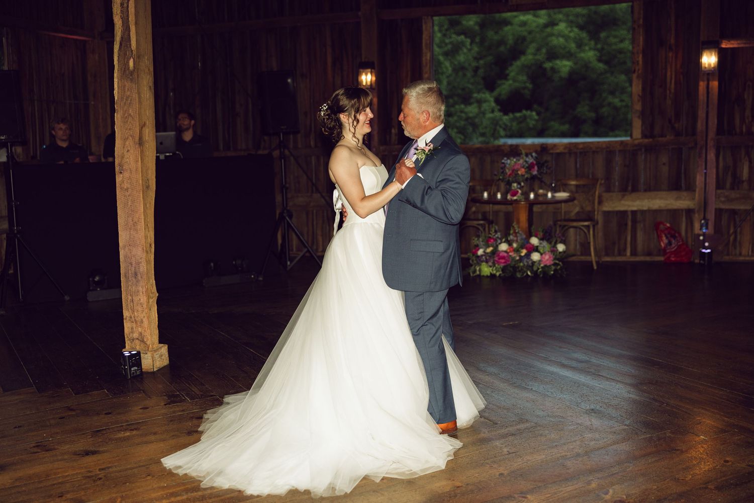 Bride in a flowing white gown shares a first dance with an older man in a gray suit inside a rustic wooden barn venue.