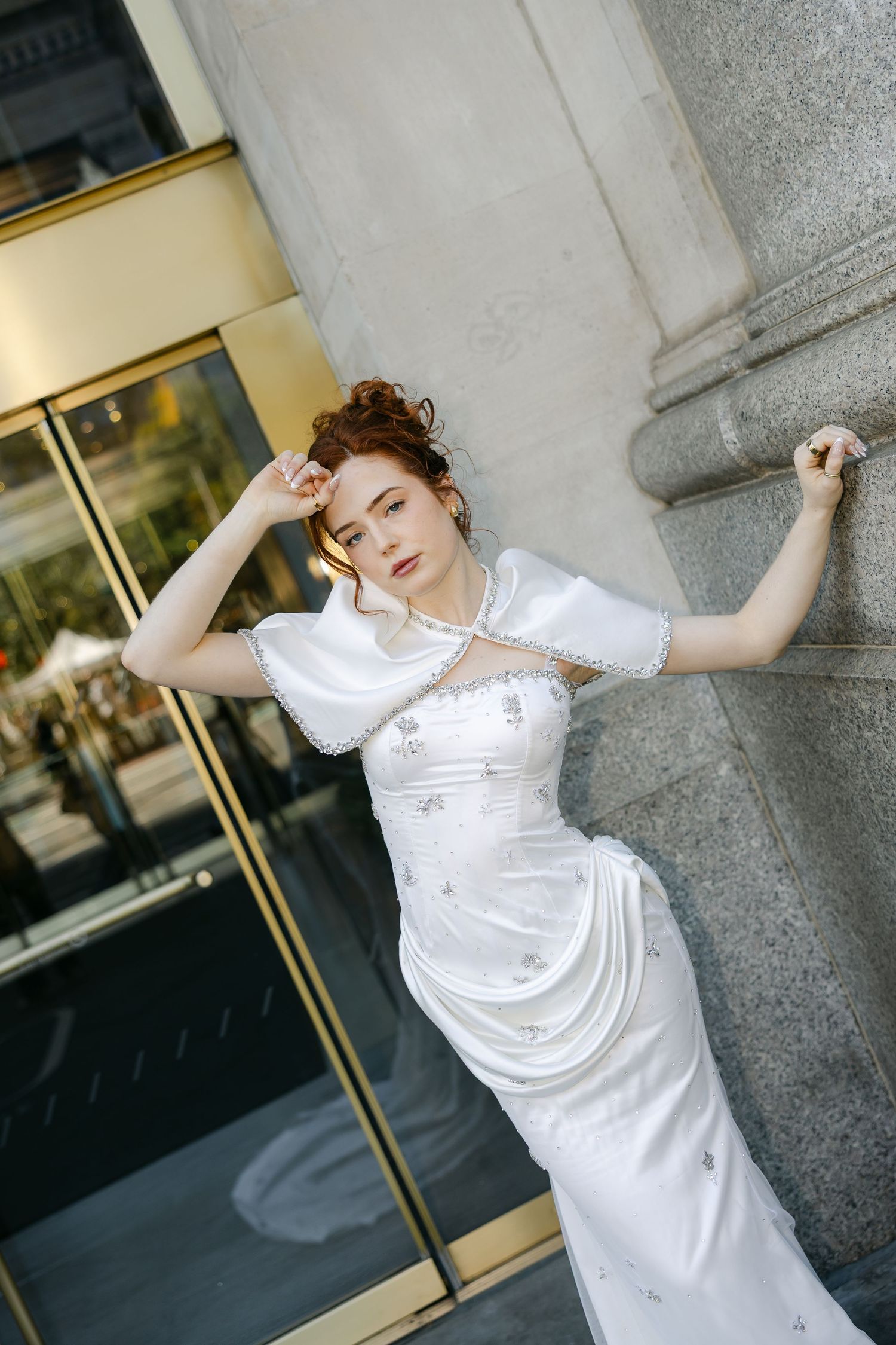 Bride with an upswept updo leans against a stone building in a fitted satin wedding gown with a sheer white shoulder cape.