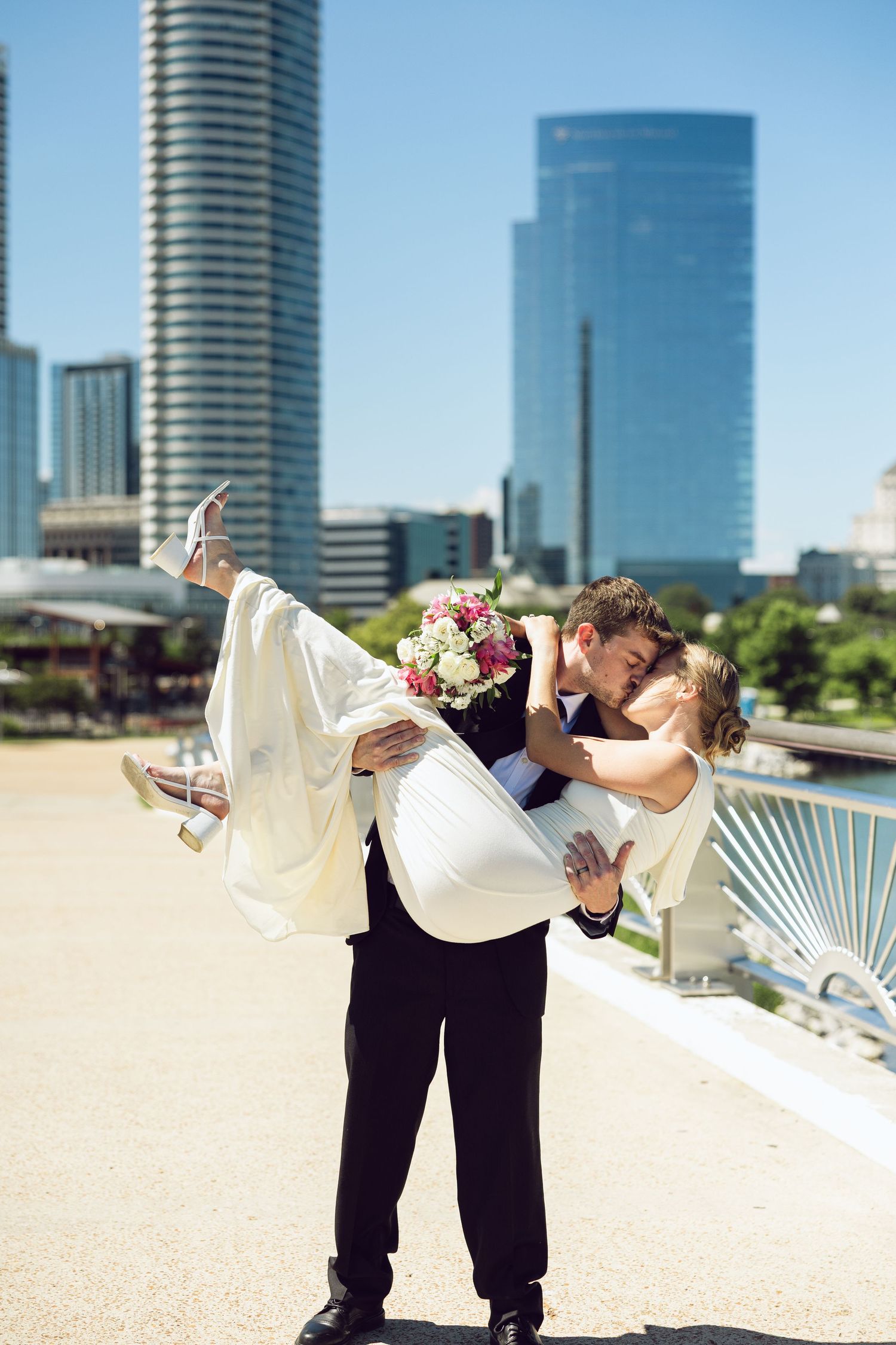 A groom lifts his bride in his arms for a kiss on a sunny city bridge with tall glass skyscrapers behind them.