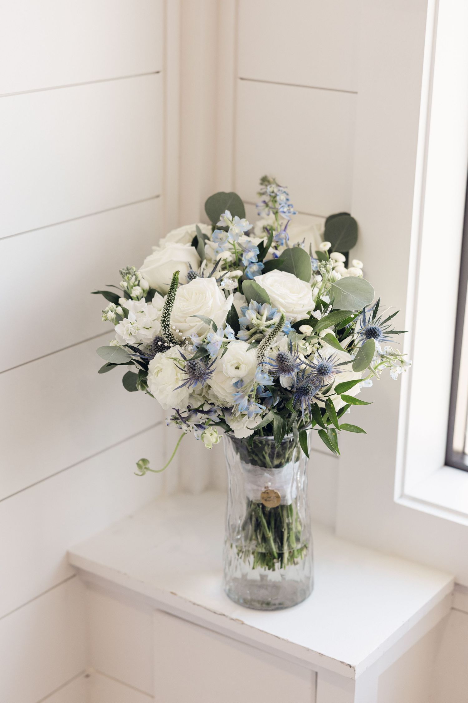 A bridal bouquet of white roses, blue thistle, and eucalyptus sits in a glass vase on a windowsill beside white shiplap walls.