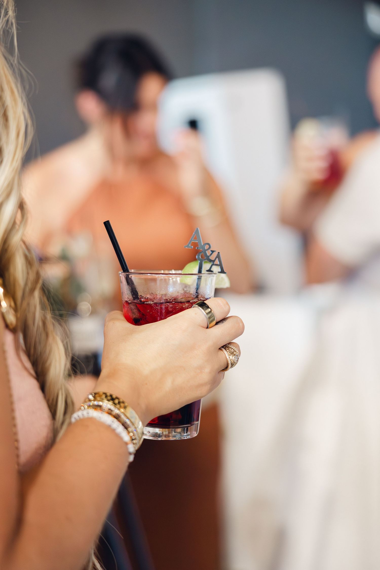 Guest in a blue lace dress holds a red cocktail with a black straw and decorative stirrer during a wedding reception.