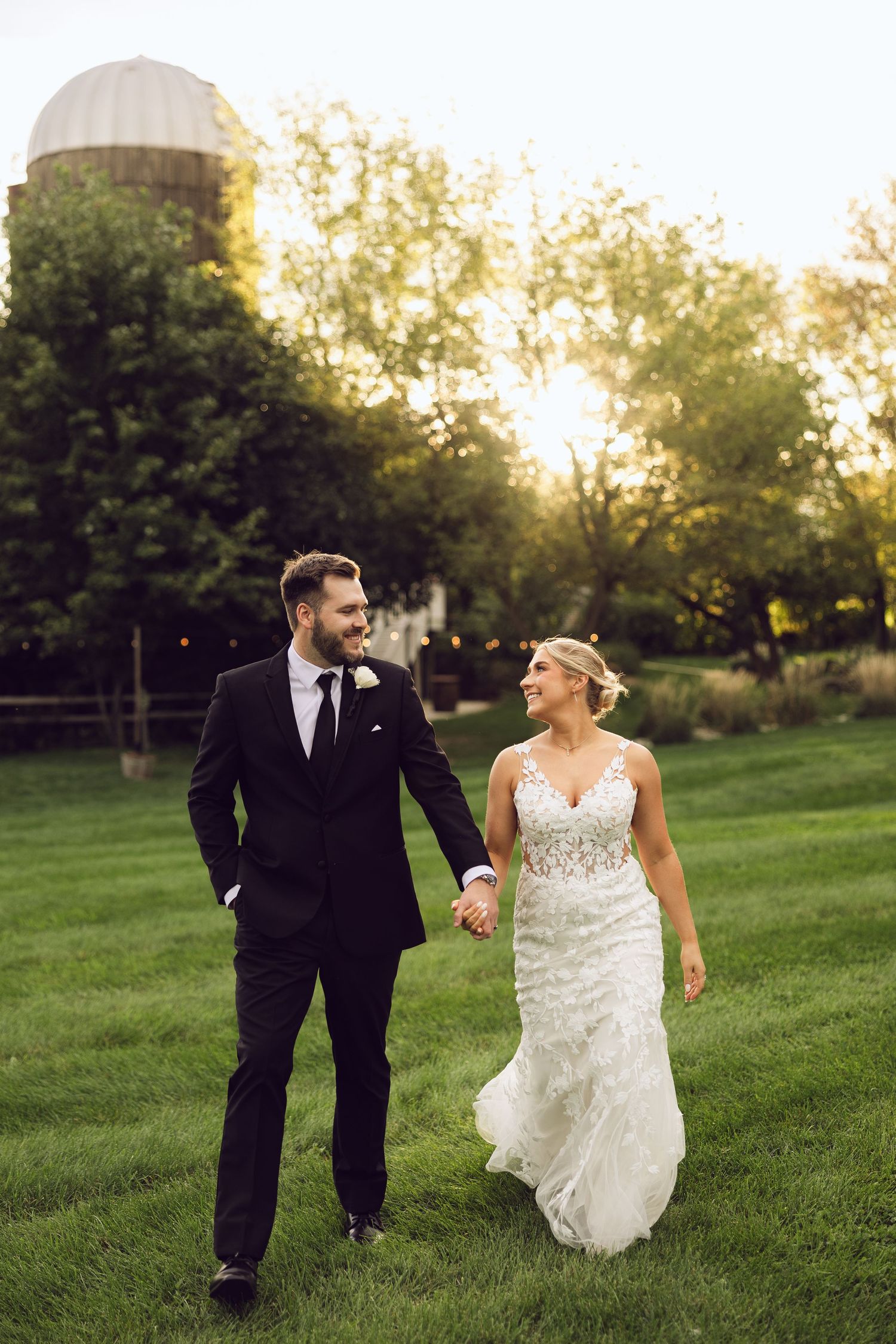 A bride in a lace wedding gown and groom in a dark suit hold hands and walk across a sunlit lawn near a silo.