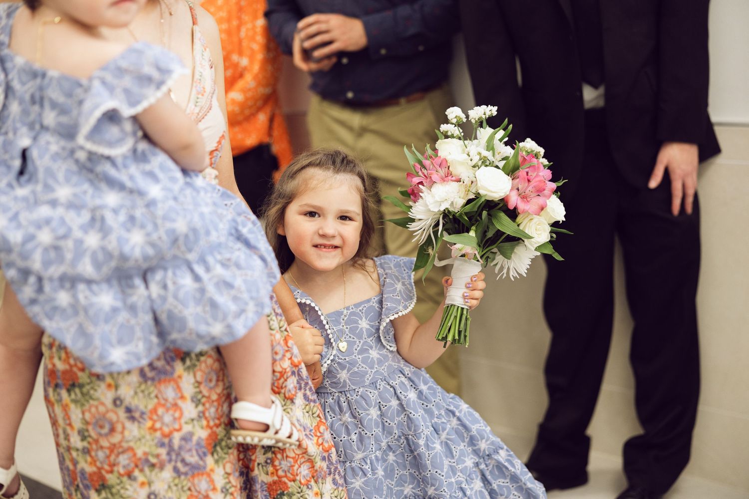 A smiling little girl in a blue floral dress holds a small bouquet of pink and white flowers at a wedding celebration.