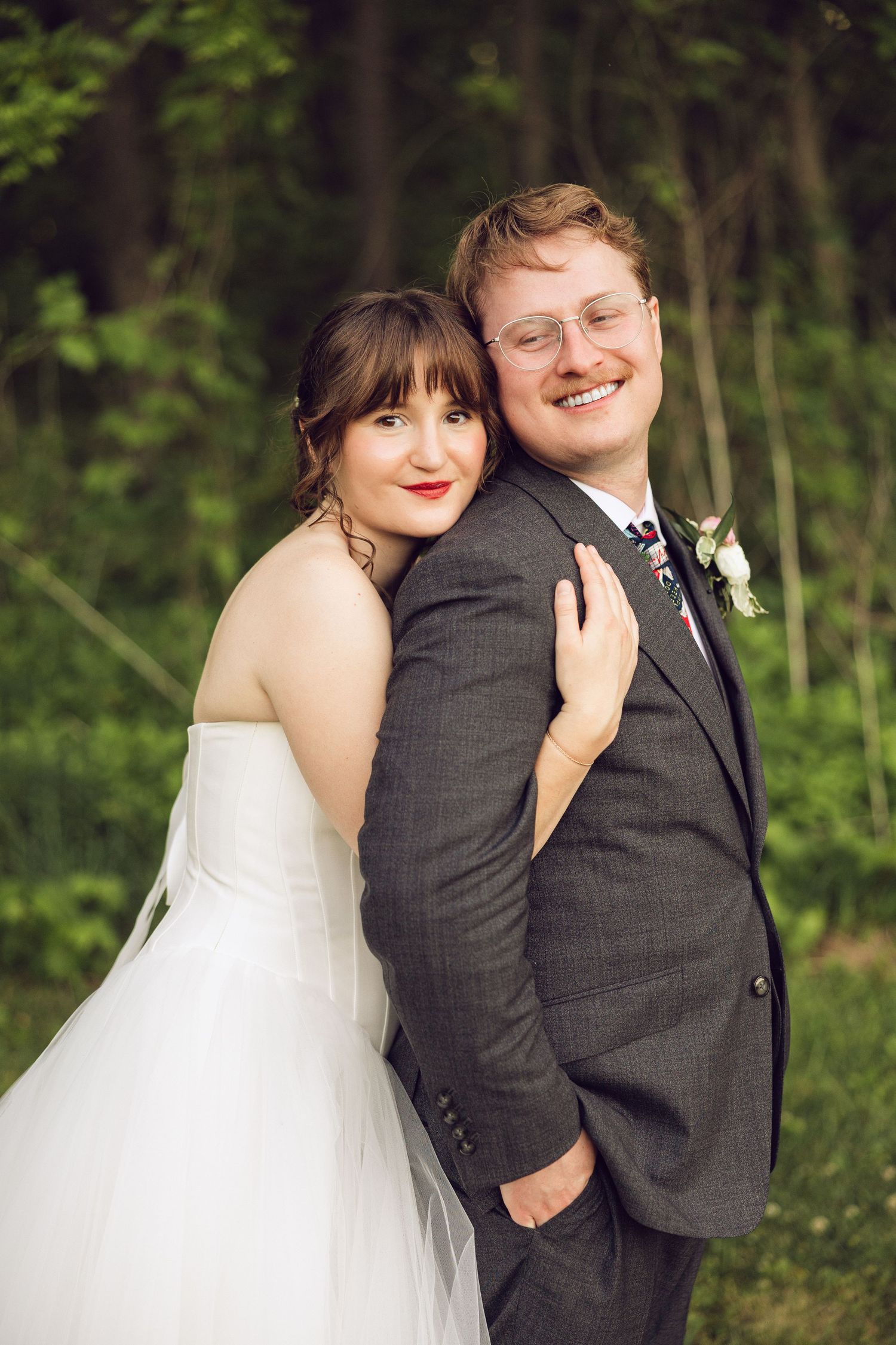 Bride in a strapless white gown hugs her smiling bespectacled groom from behind in a lush green woodland setting.