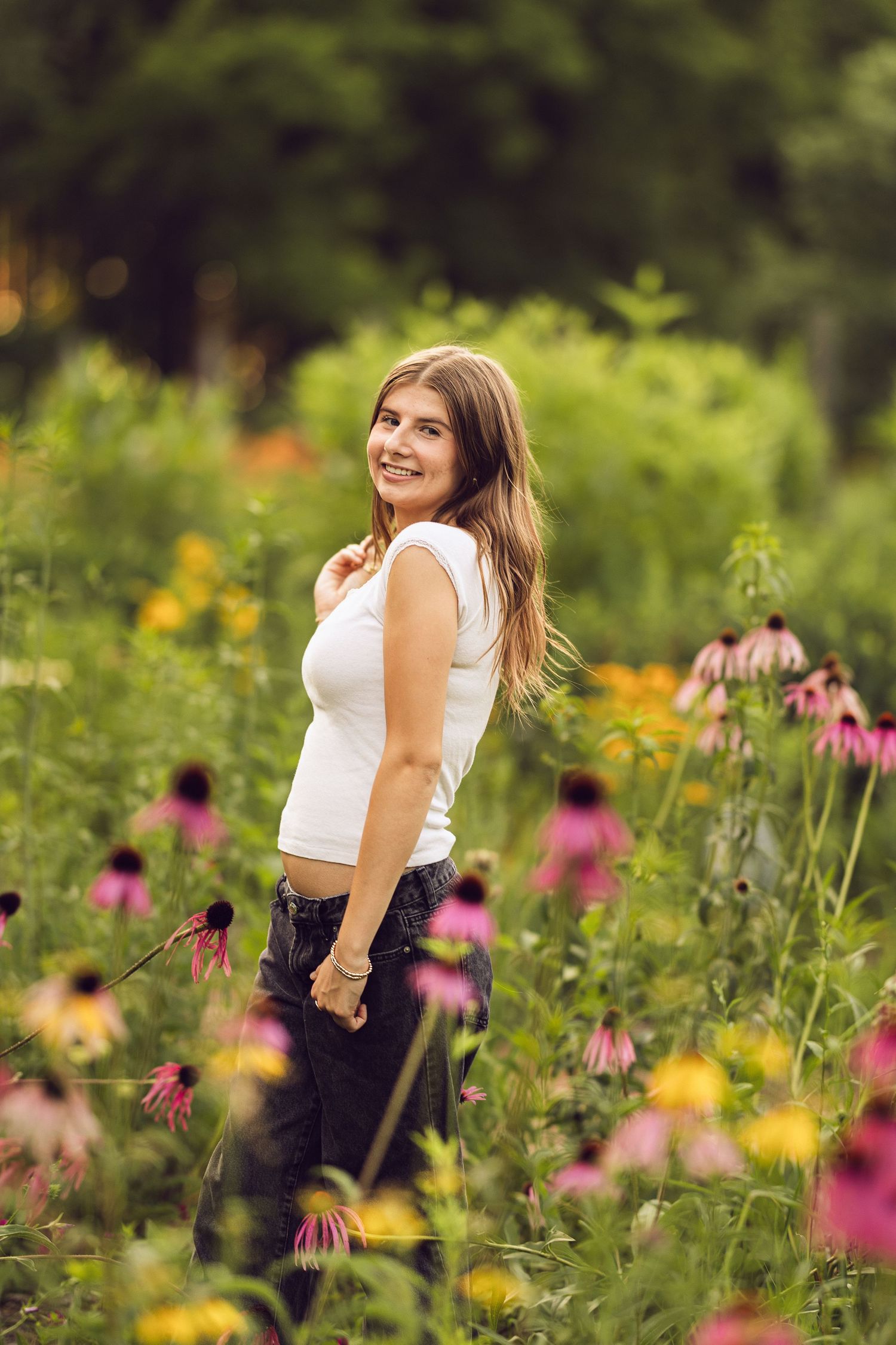 Smiling young woman in a white tank top and dark jeans poses among pink coneflowers in a sunlit summer meadow.