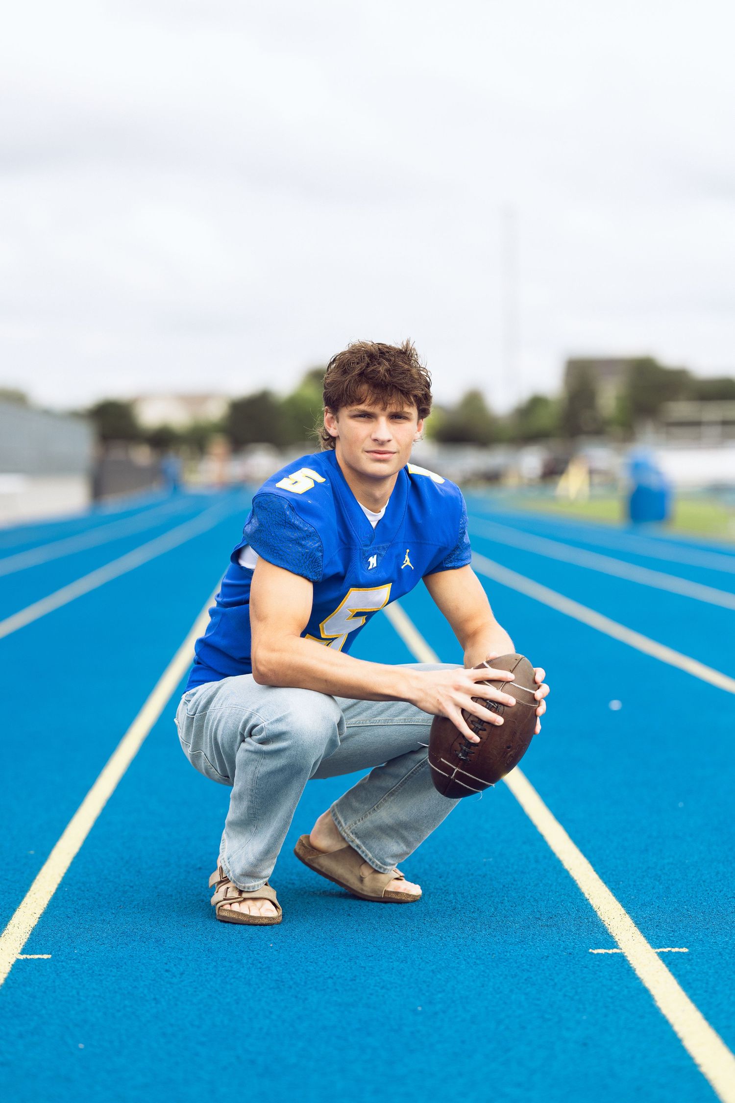 Young man in a blue football jersey crouches on a blue running track while holding a football for a senior portrait.