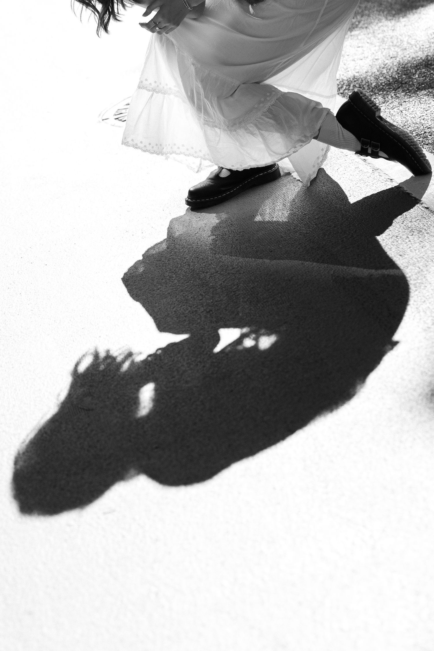 Black and white close-up of a girl in a lace-trimmed dress and black shoes casting a long dramatic shadow on pavement.