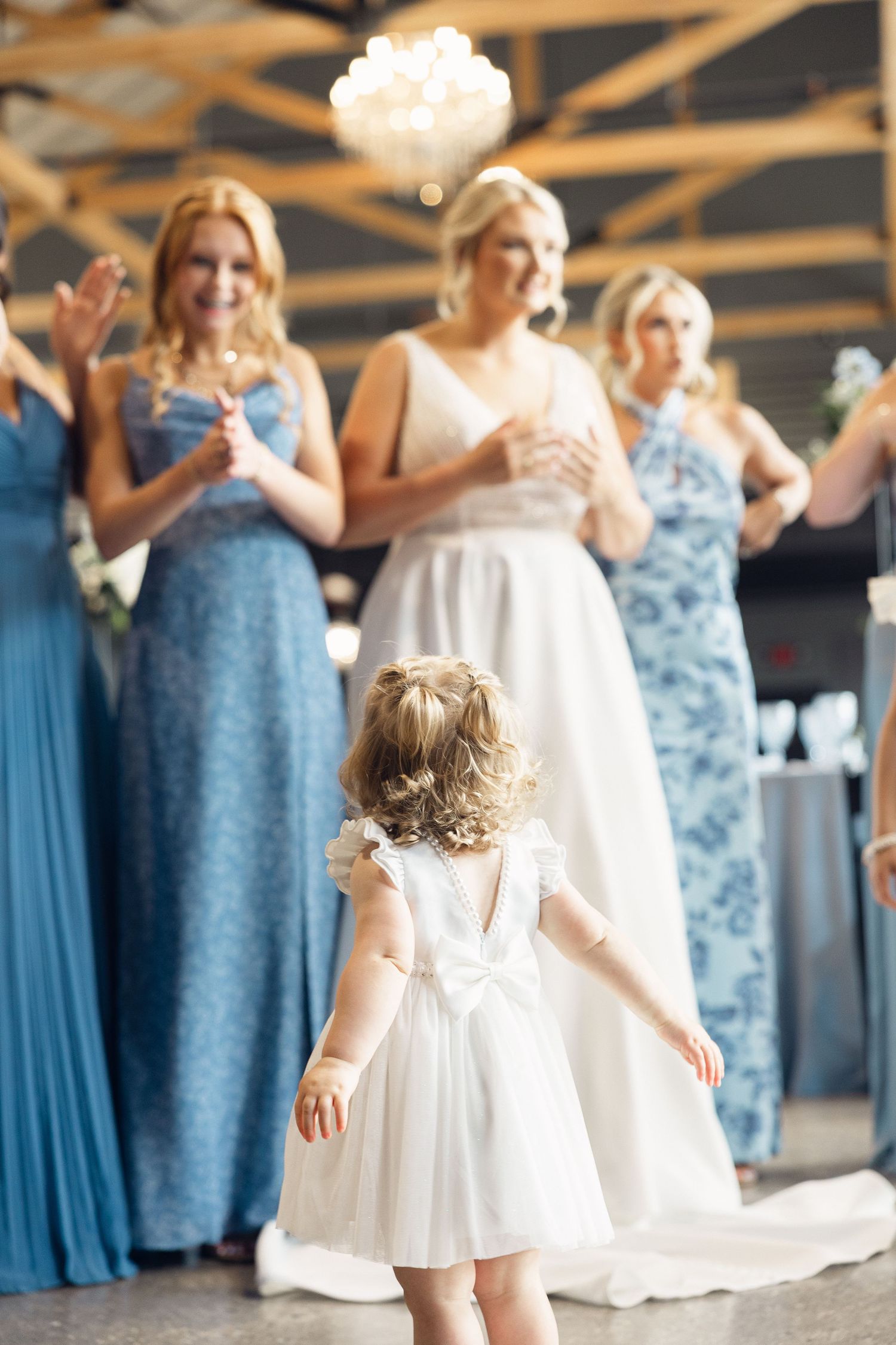 A flower girl in a white dress faces bridesmaids in blue gowns and a smiling bride beneath a chandelier at a wedding reception.