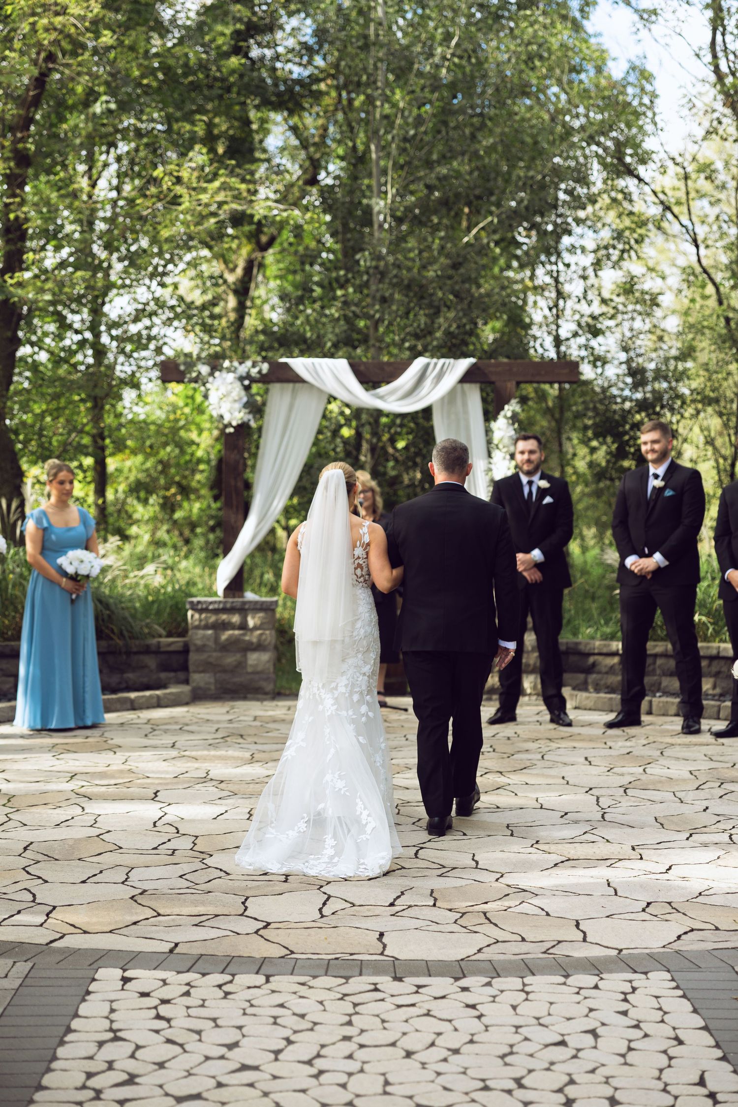 A bride in a lace gown and long veil walks toward a draped wedding arch with groomsmen and a bridesmaid waiting outdoors.