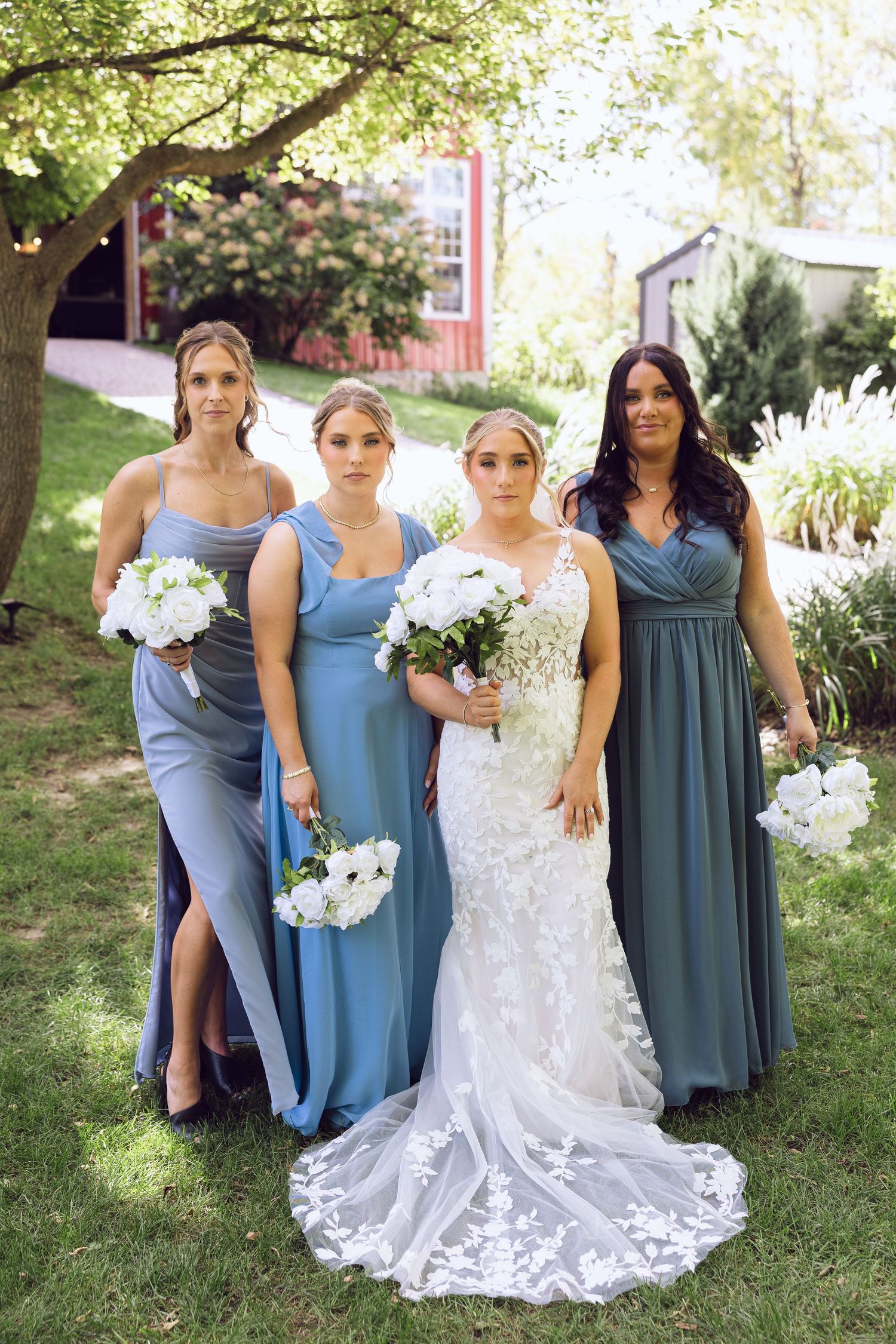 Bride in a lace gown stands among three bridesmaids in blue dresses, all holding white bouquets on a sunny lawn.