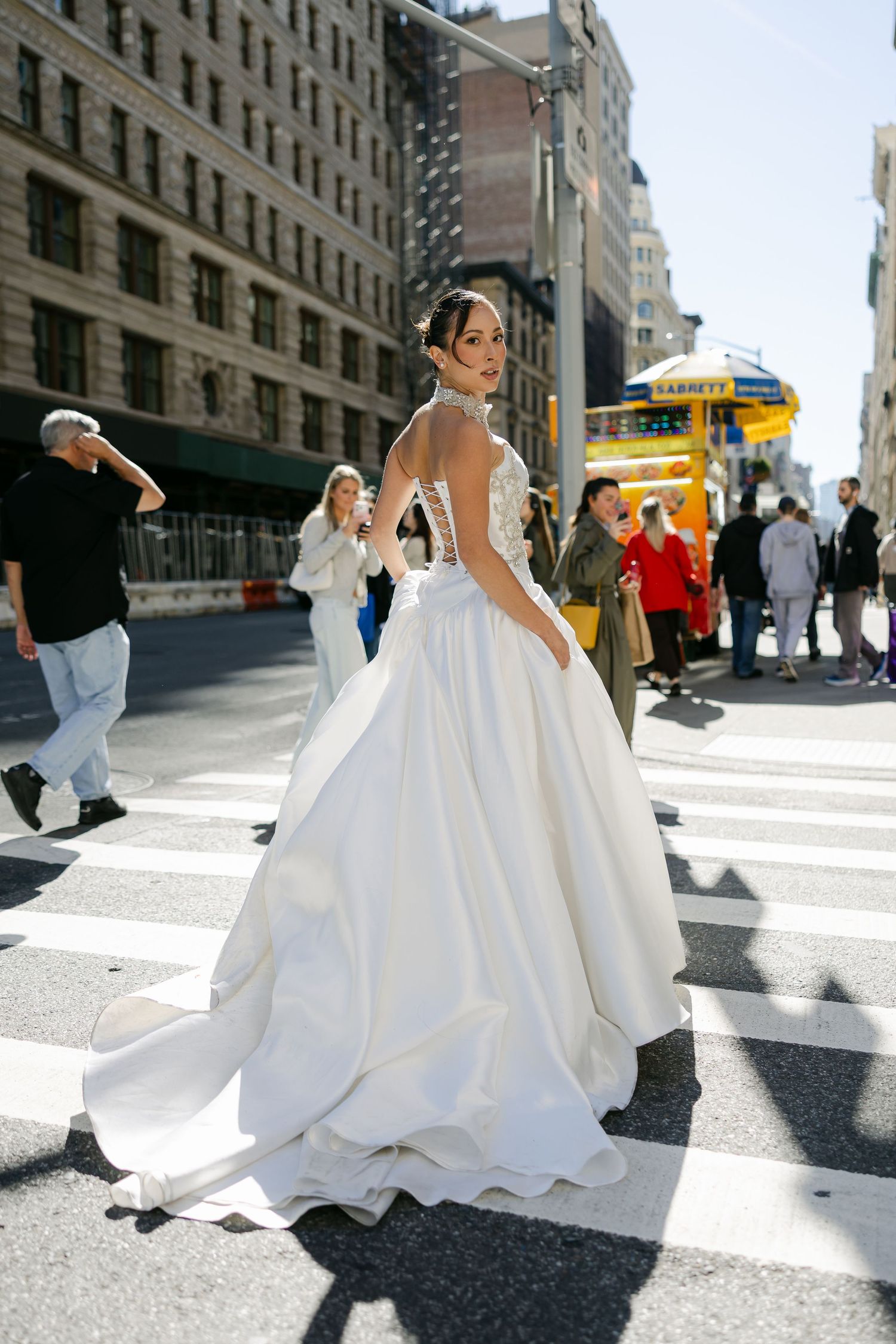 Bride in a strapless satin ball gown with a corset bodice stands in a sunny New York City crosswalk as pedestrians pass behind.