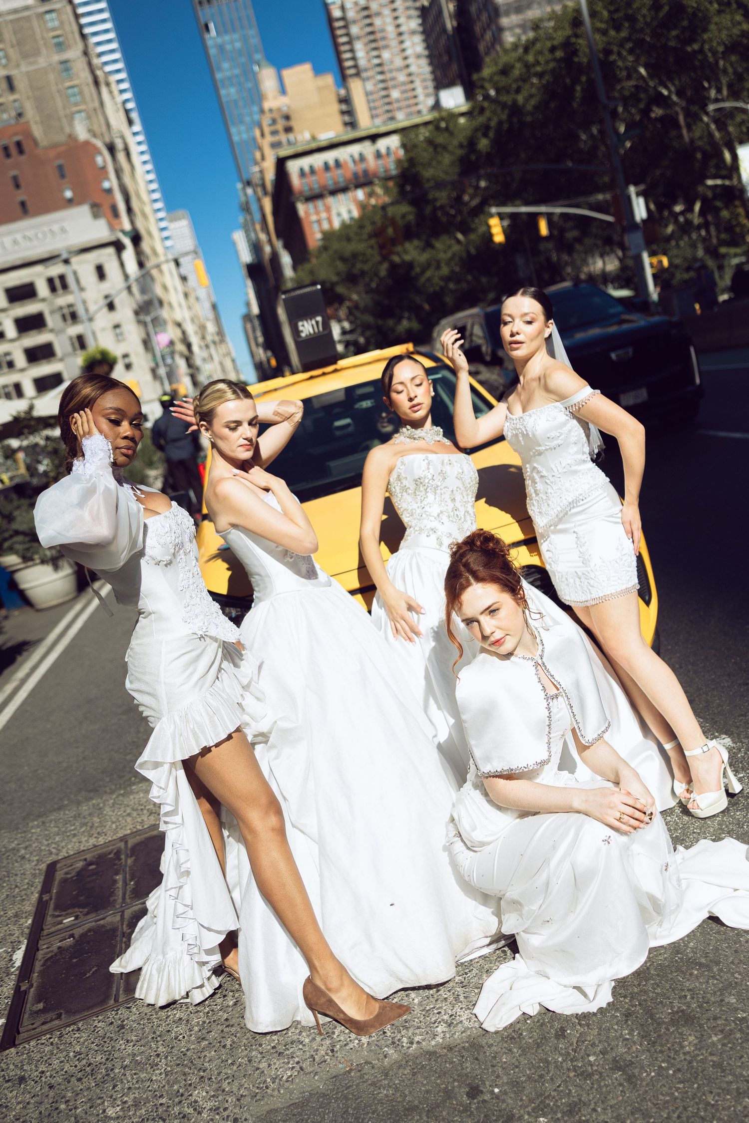 Models in elegant white bridal dresses pose around a yellow taxi on a sunlit New York City street.