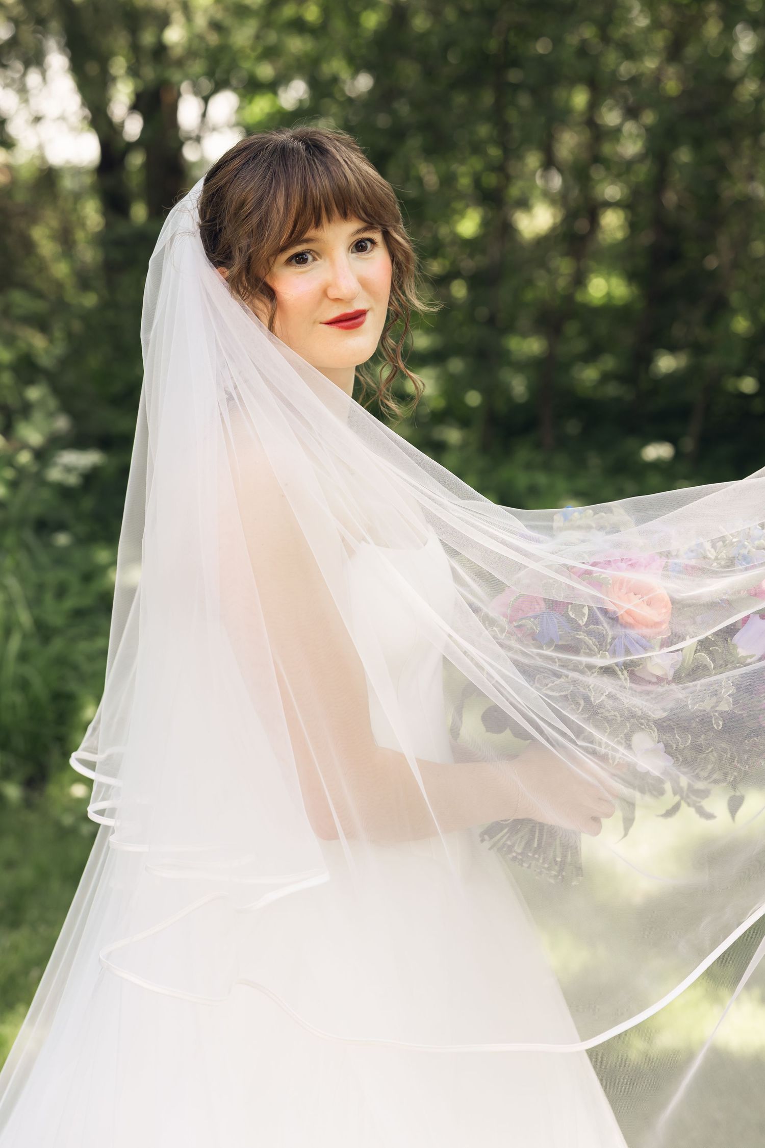 Smiling bride with bangs and red lipstick stands outdoors in a flowing tulle veil, holding a colorful wedding bouquet.