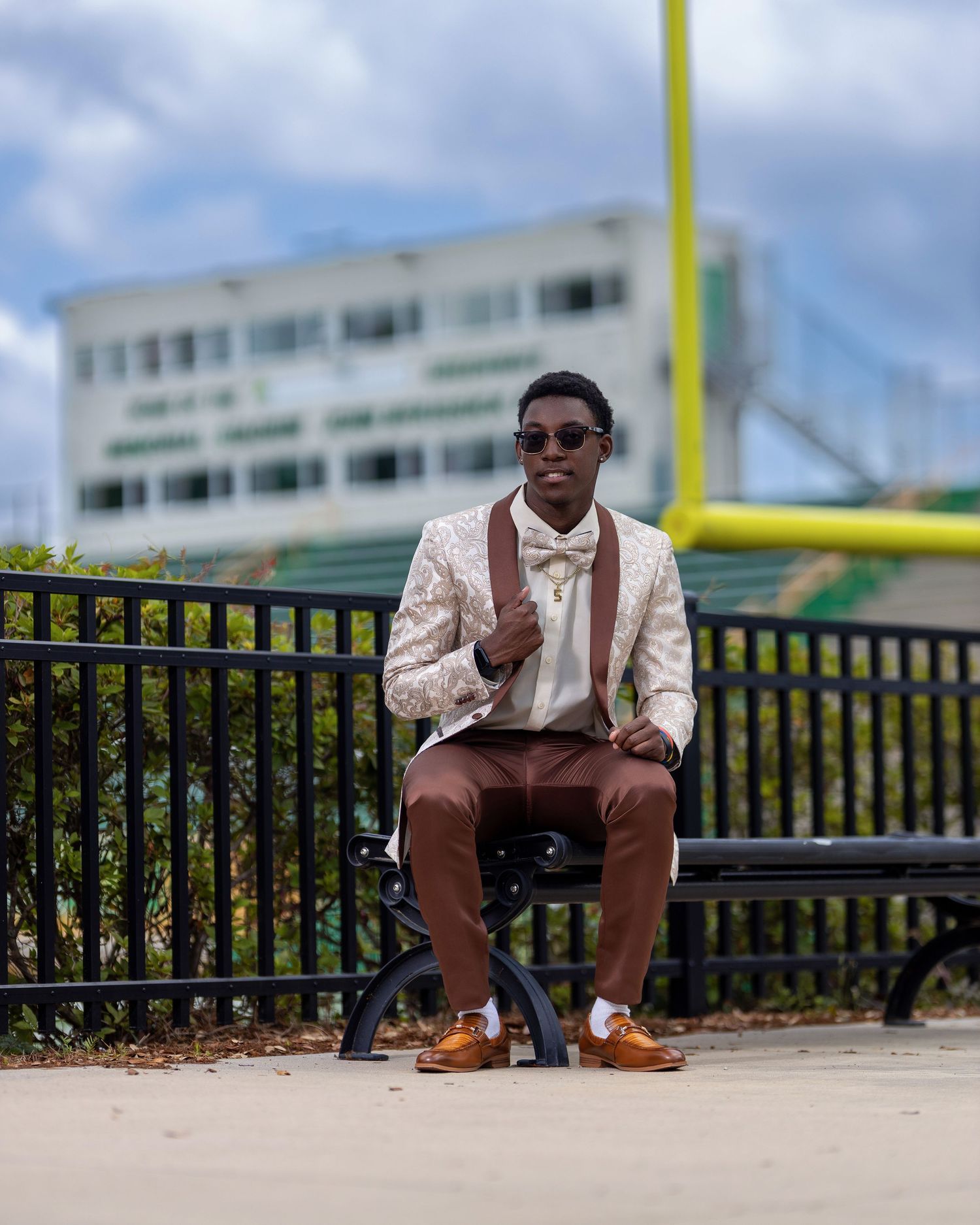 High school senior in a patterned jacket and bow tie seated near a football field