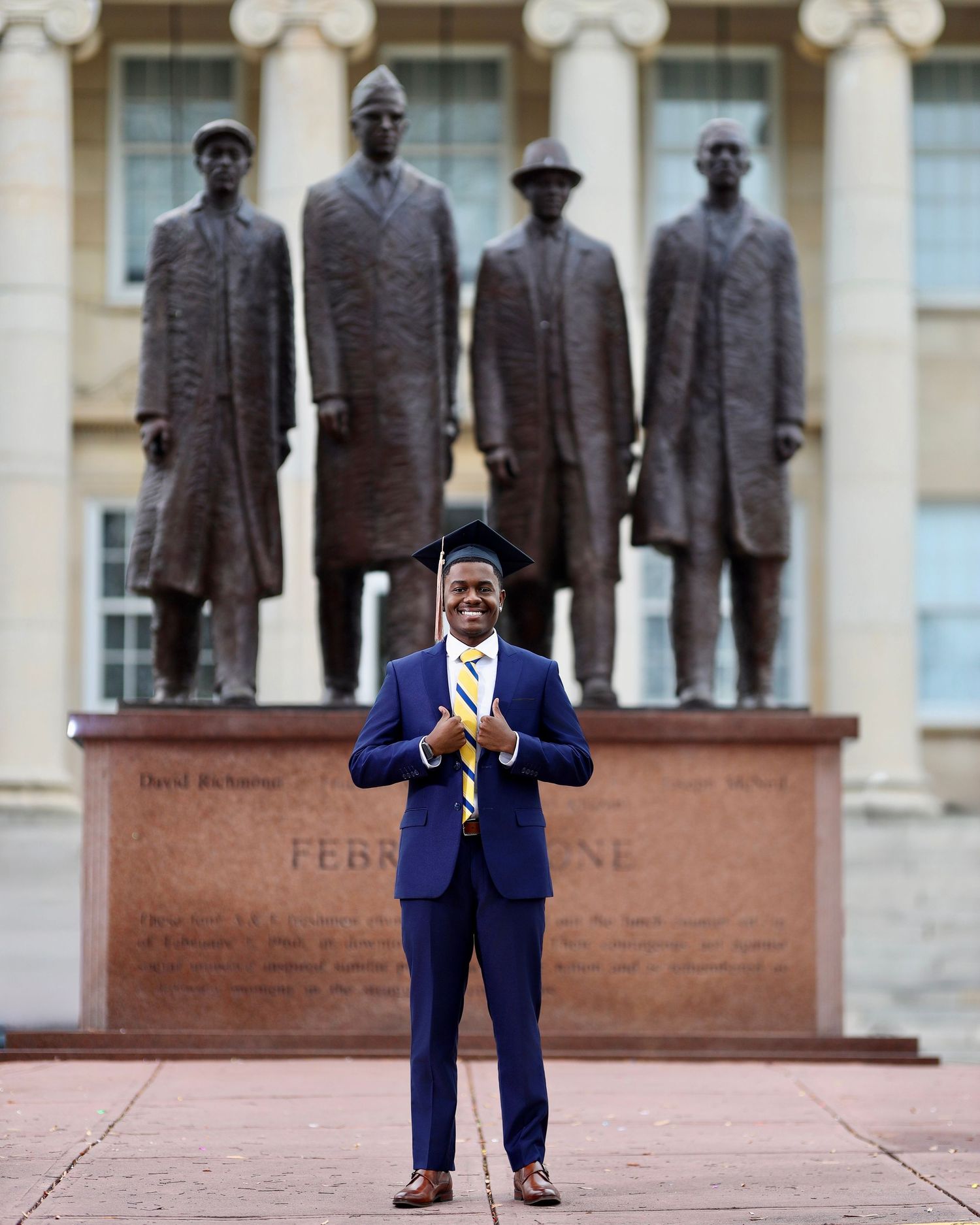Graduate in a blue suit and cap posing in front of the February One statues