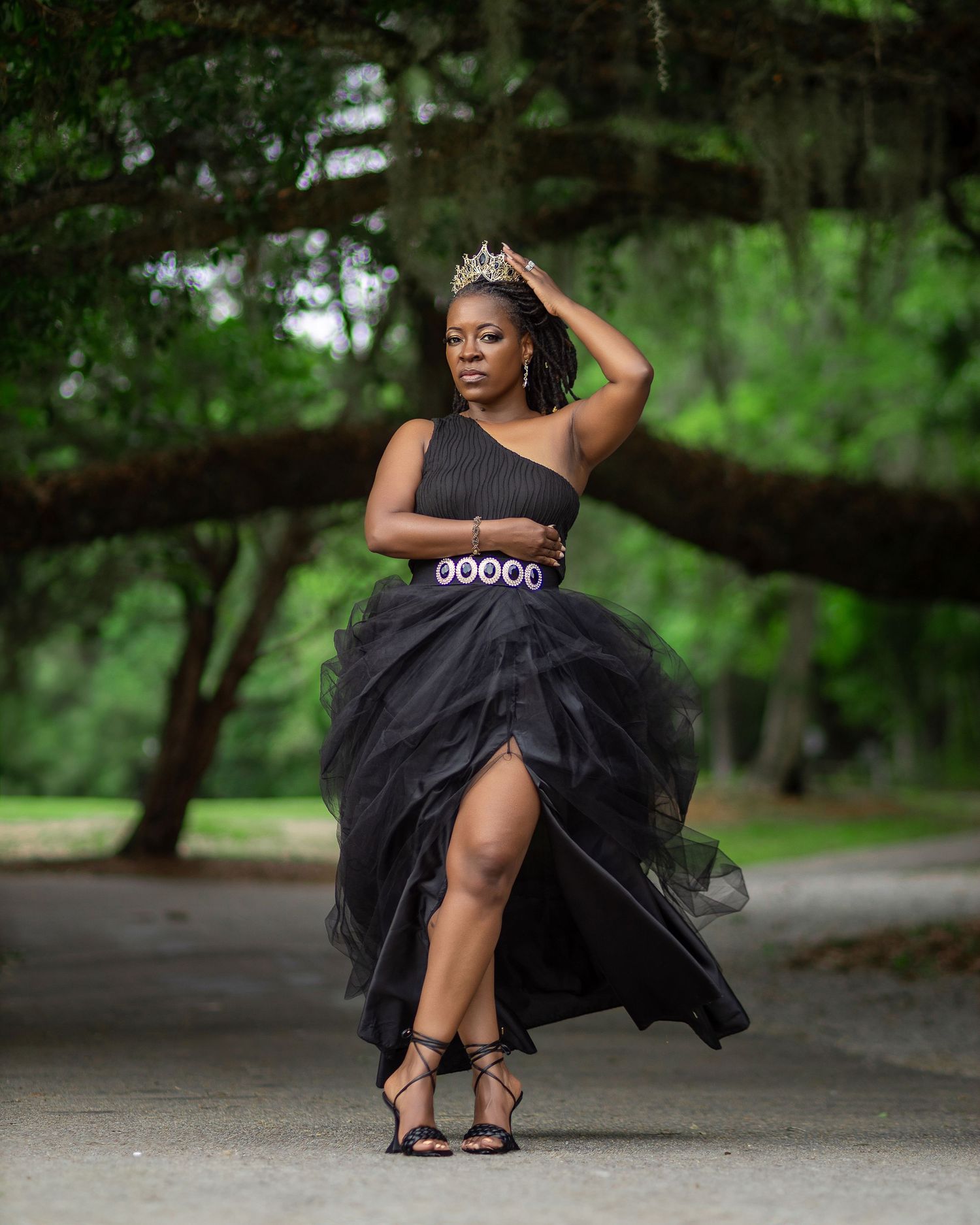 Woman wearing a crown and black gown posing on a tree-lined path