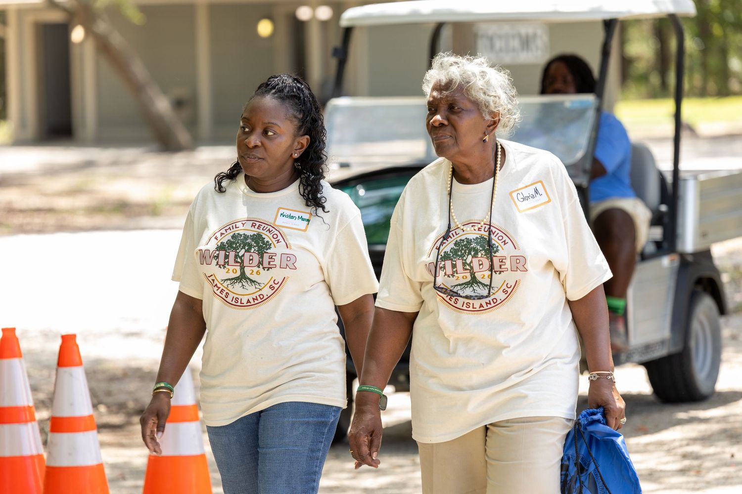 Two women in matching reunion t-shirts walk hand-in-hand at an outdoor family reunion event.