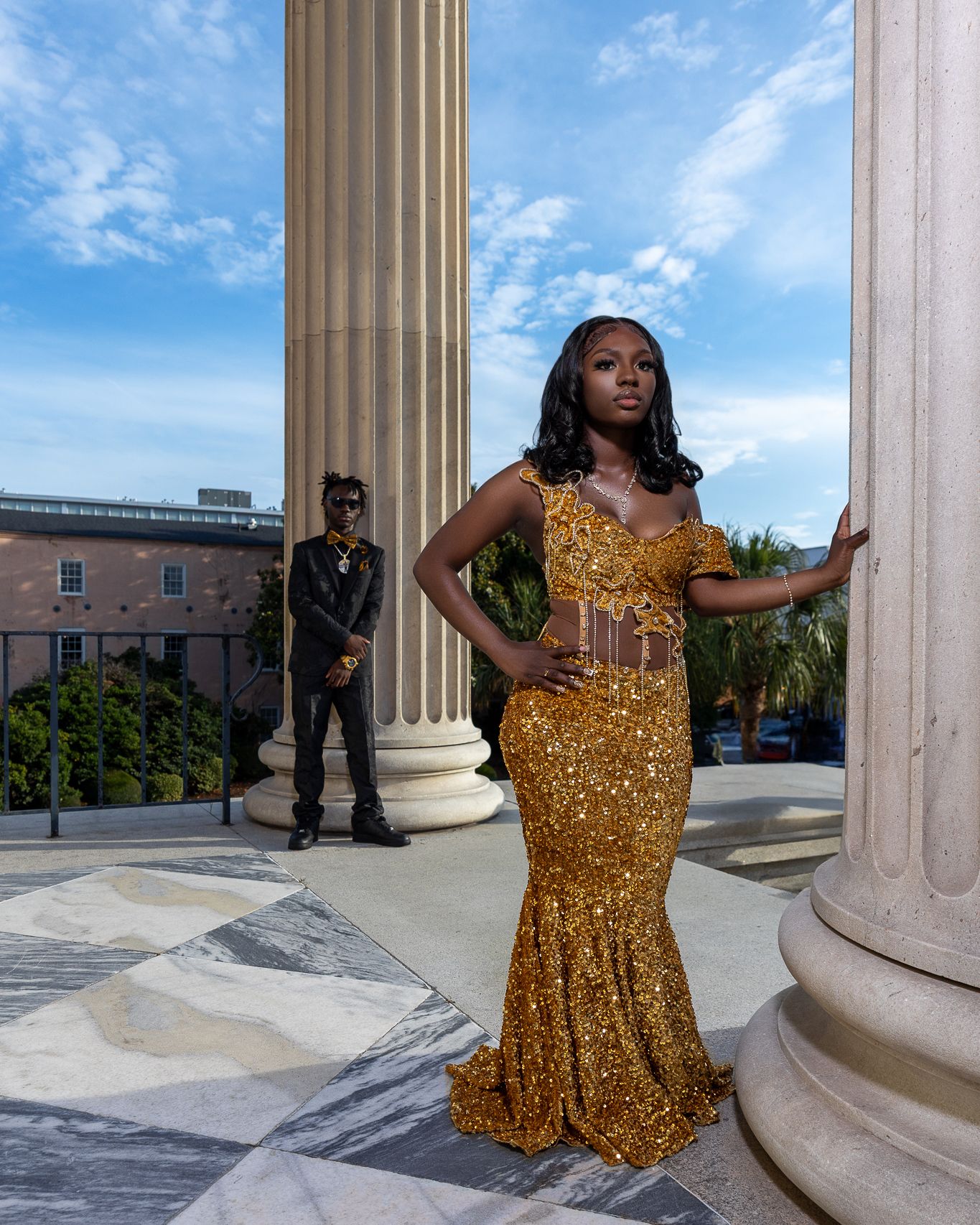 Woman in gold sequin mermaid gown poses gracefully between classical columns at a grand building.