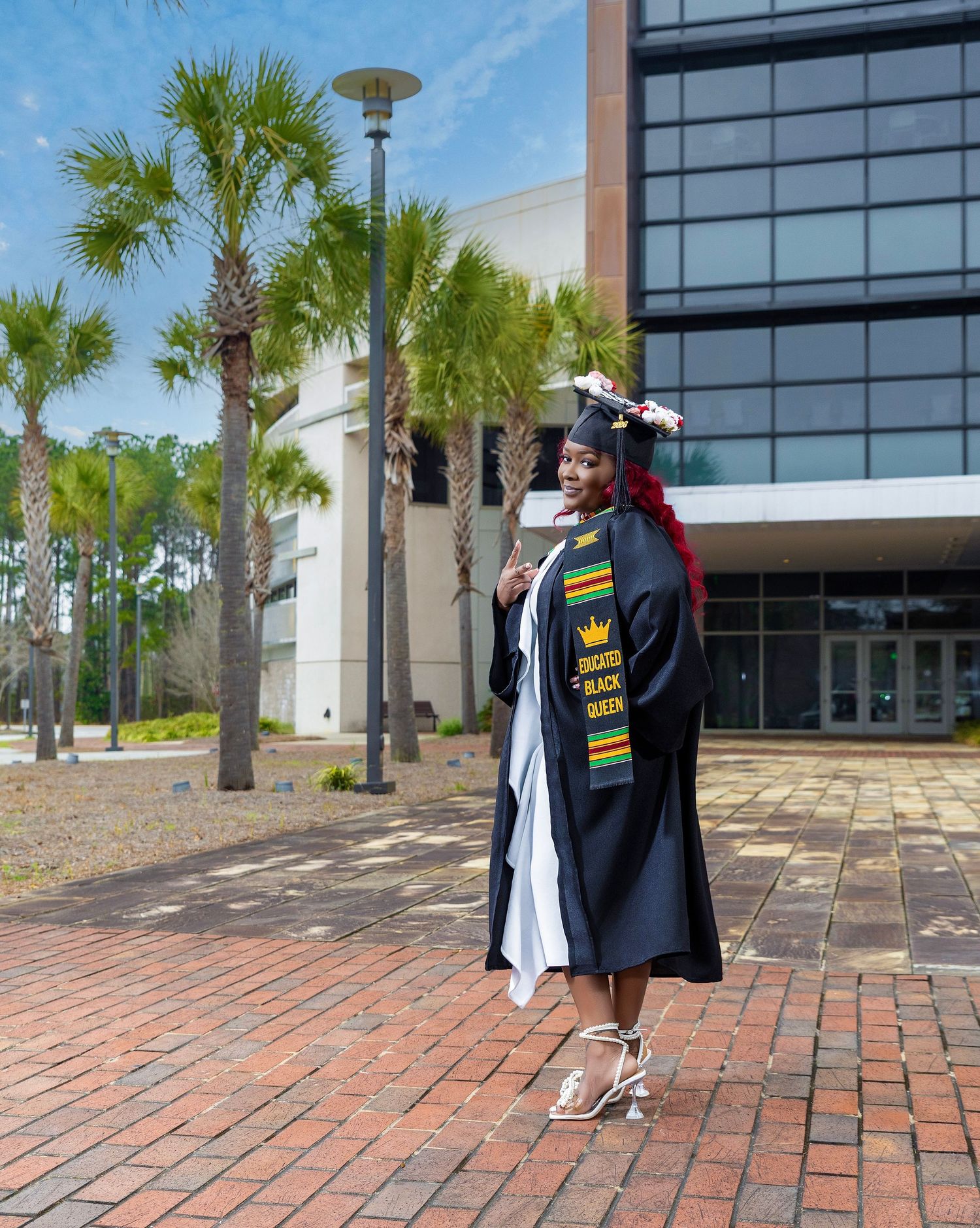 College graduate wearing a cap and gown standing outside a campus building lined with palm trees