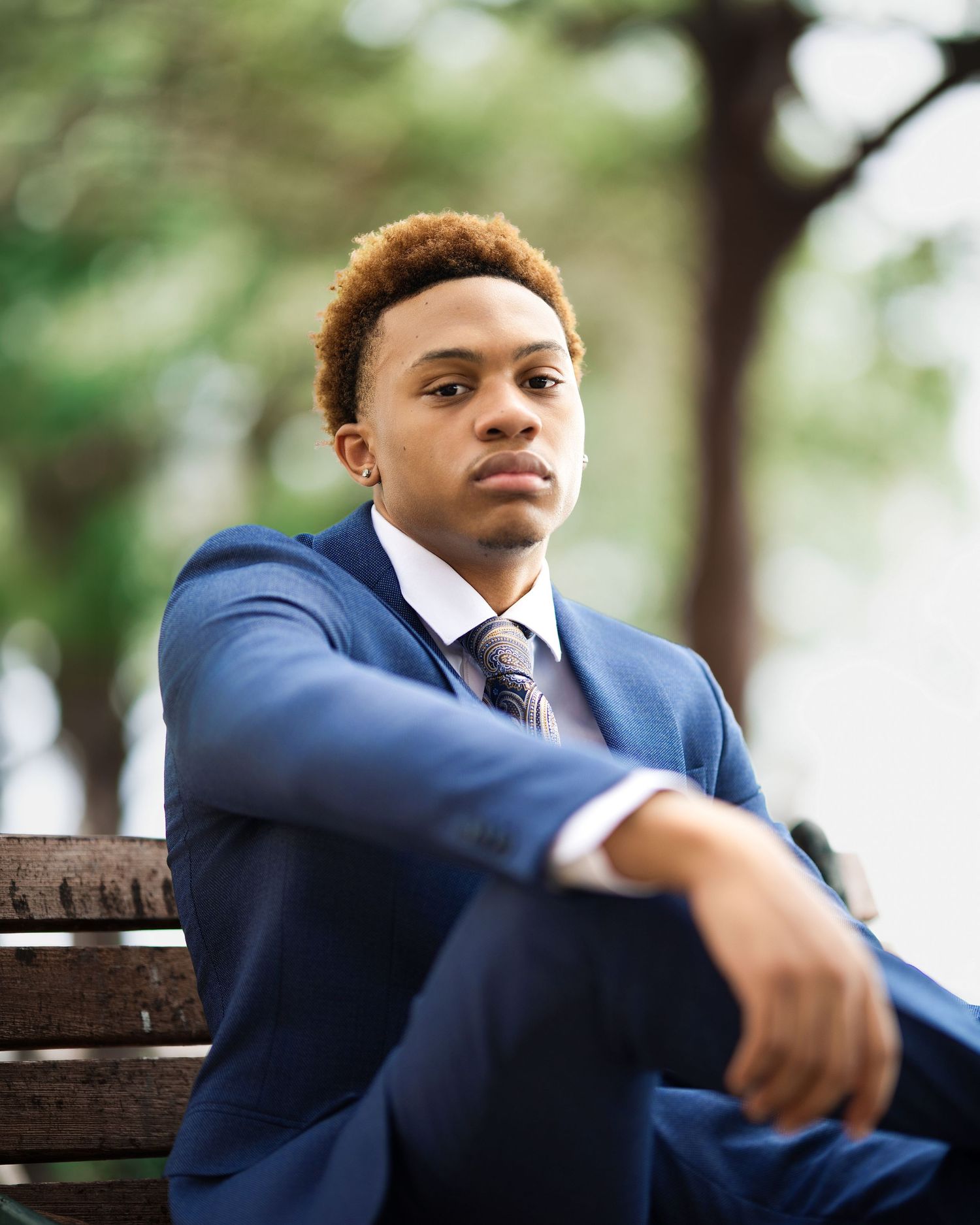 Young man in blue suit sits thoughtfully on a bench in a park setting.