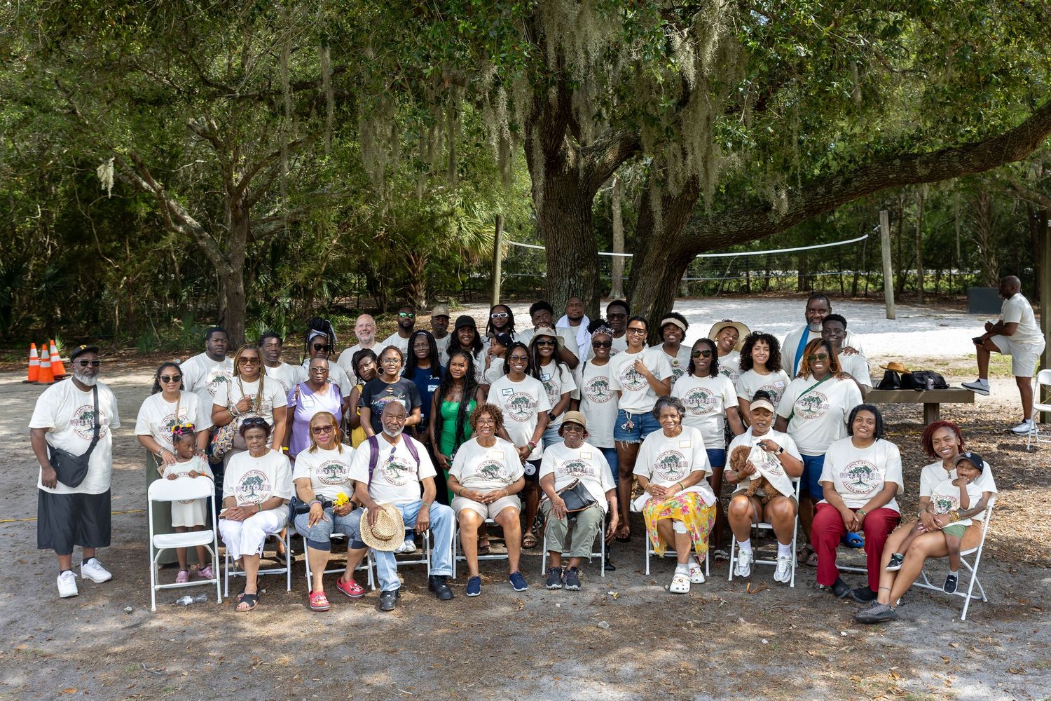 Large family reunion group photo outdoors under oak trees, all wearing matching white t-shirts.