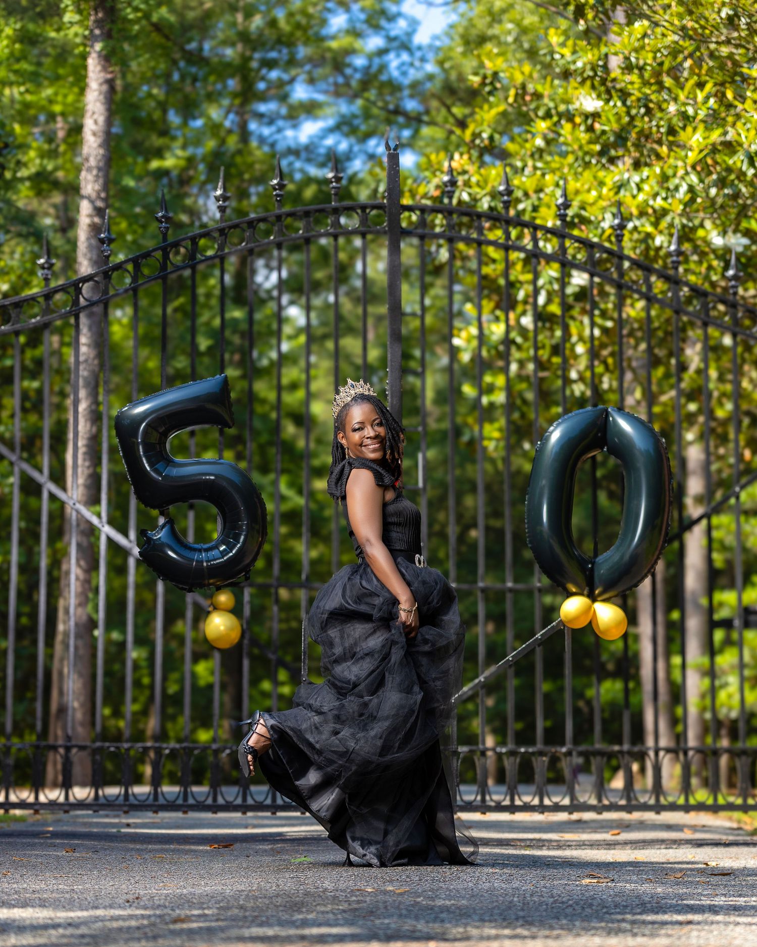Woman wearing a crown and black gown posing between large 50th birthday balloons