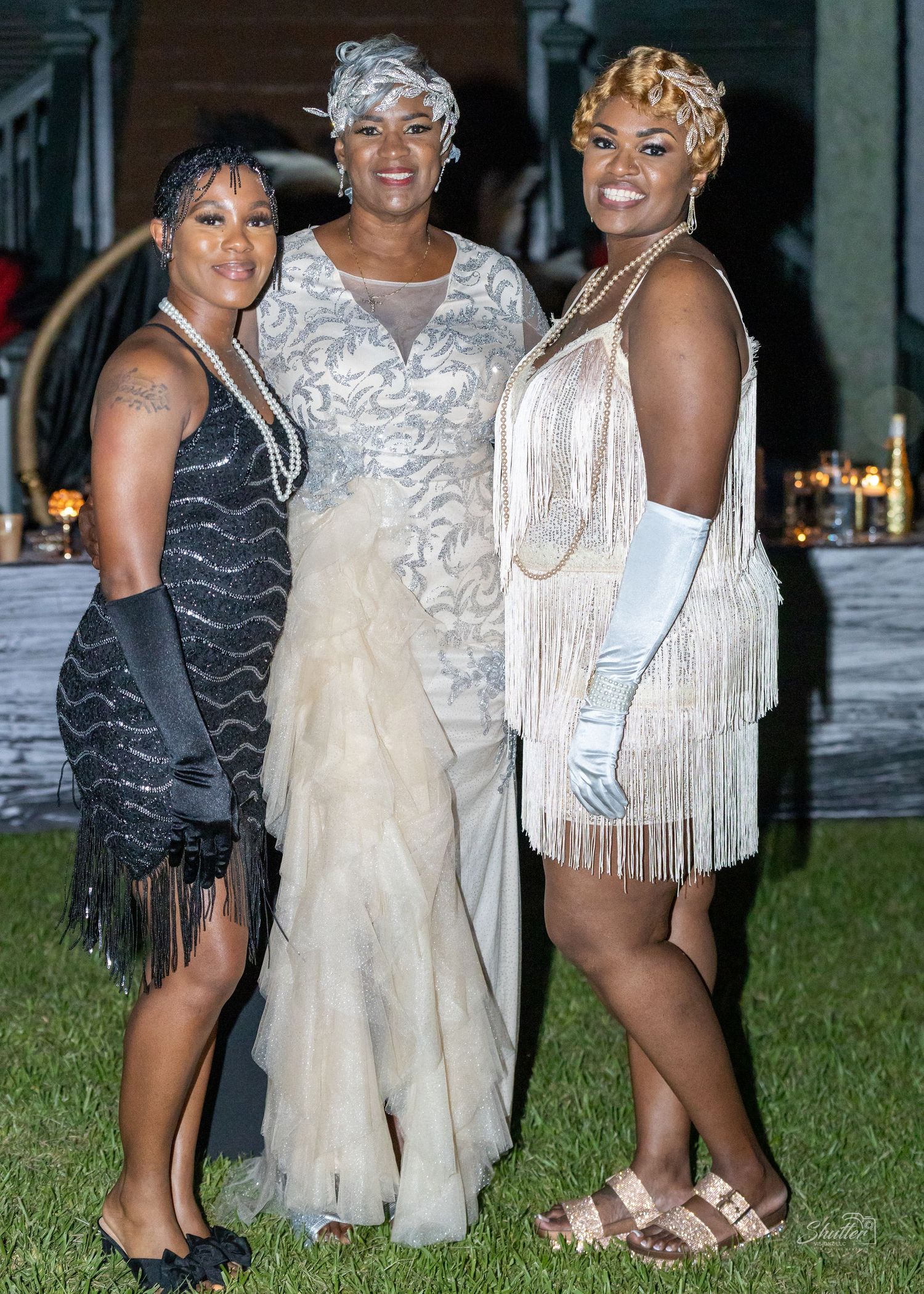 Three women in glamorous 1920s-inspired fringe dresses pose together at an outdoor evening celebration.