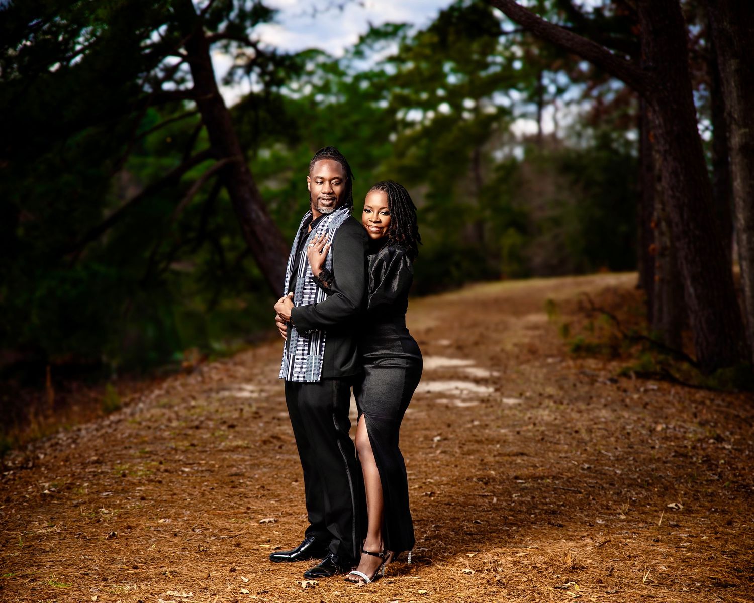 A couple poses elegantly on a forest dirt path, man in plaid shirt and black pants, woman in black slit dress.