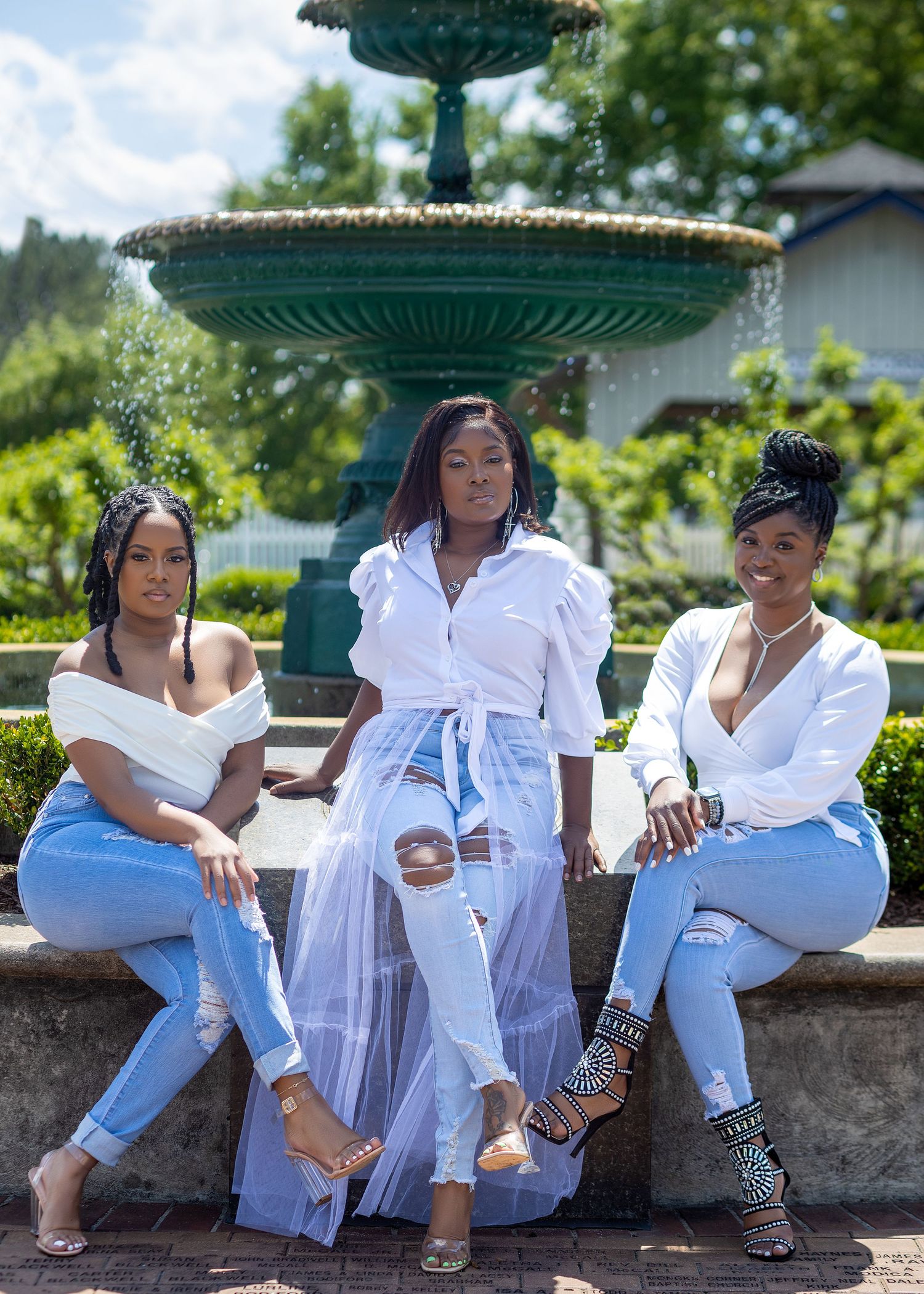 Three women posing together in white tops and jeans beside a decorative fountain