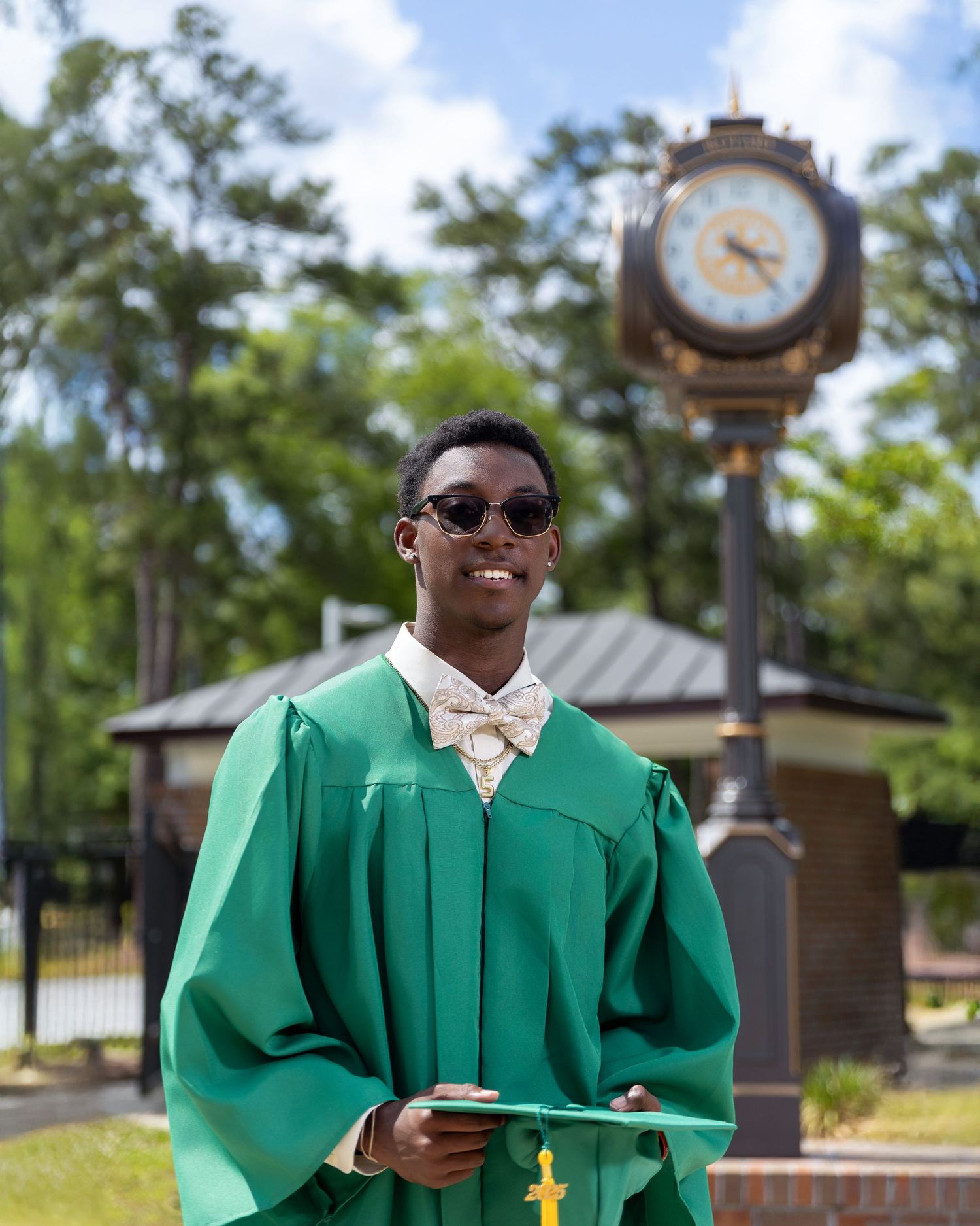 High school senior wearing a green graduation gown and holding a diploma beside an outdoor clock