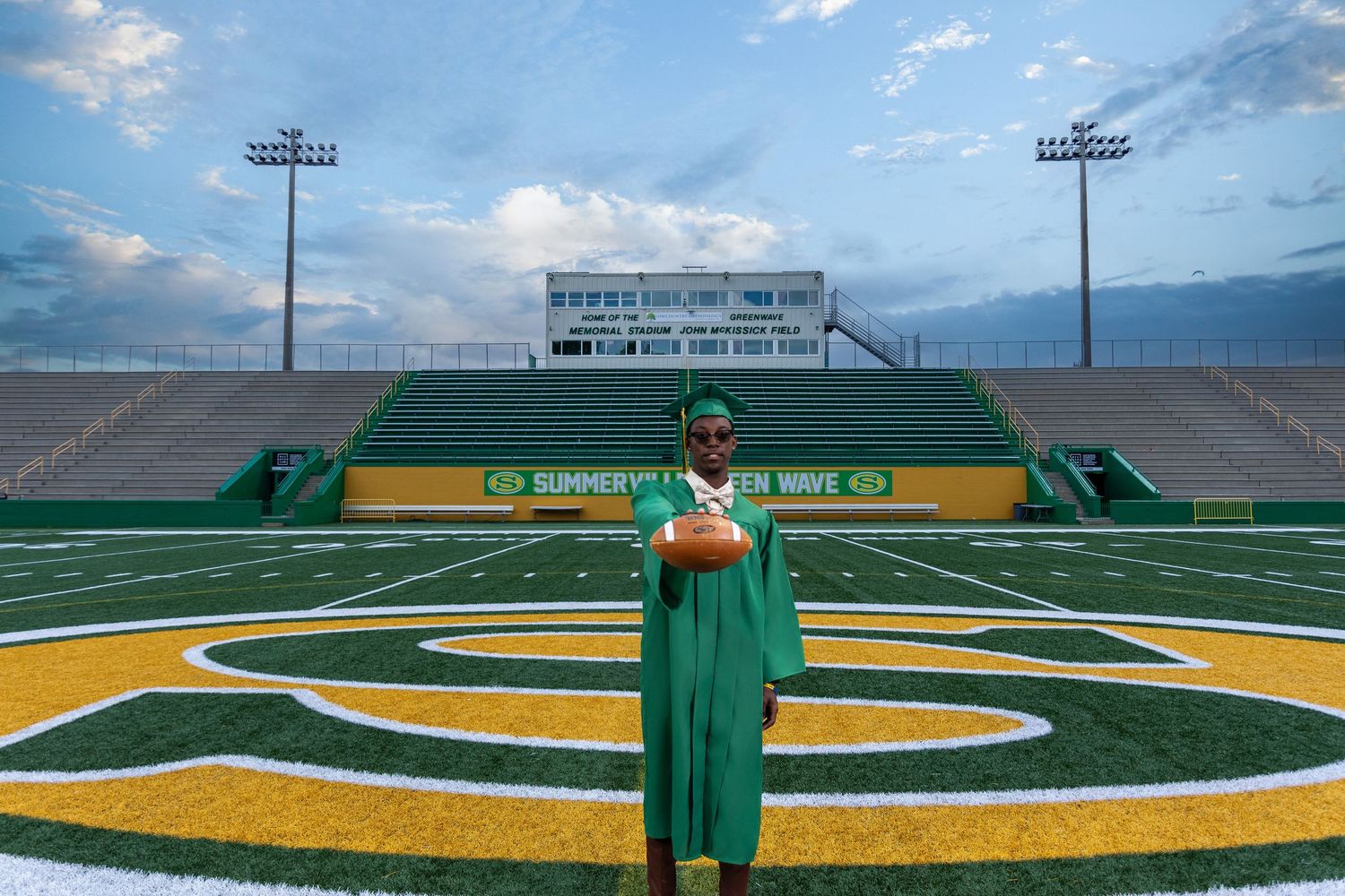 High school senior in a green graduation cap and gown holding a football on the field