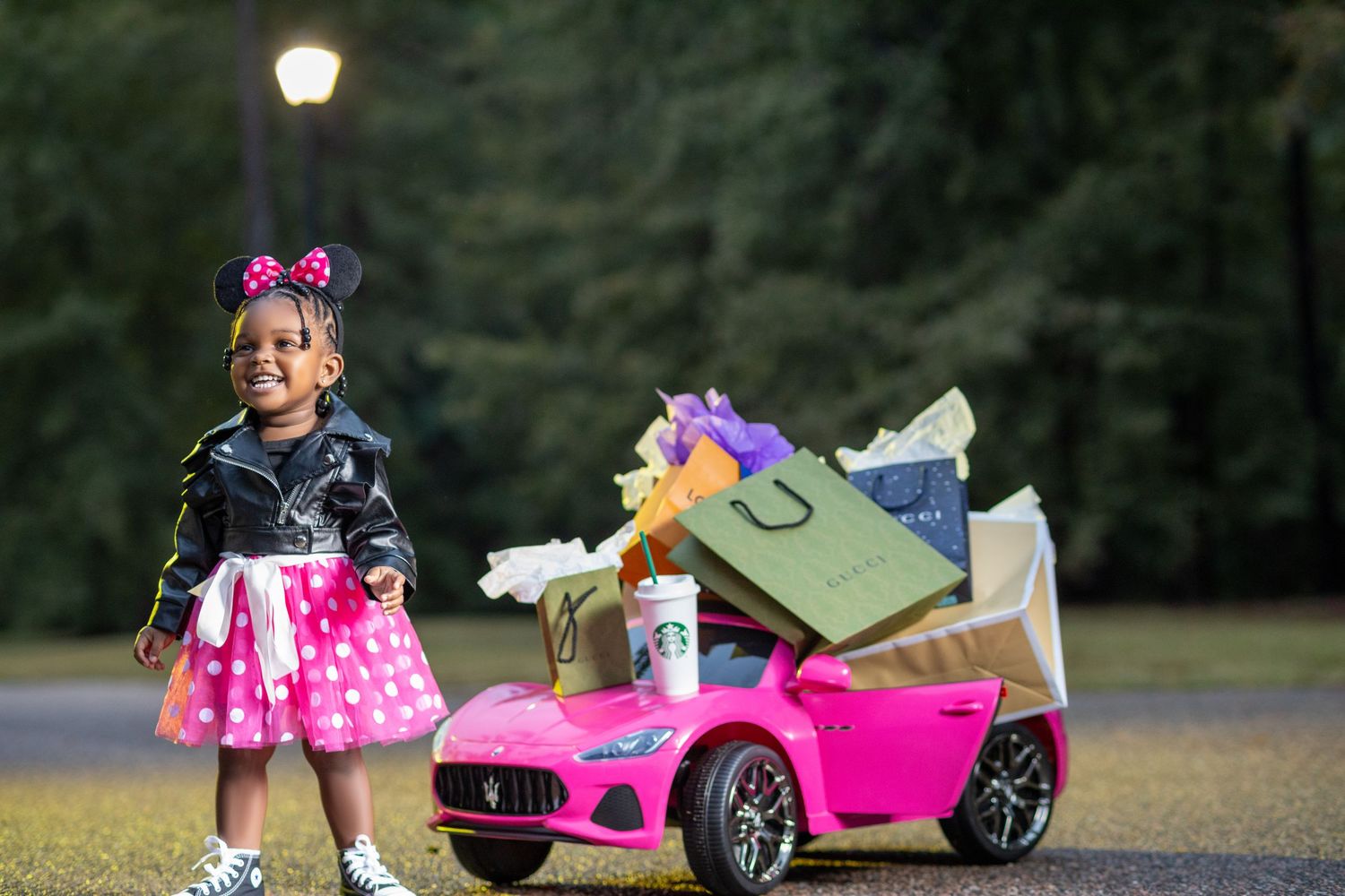 Happy girl in Minnie Mouse dress stands beside pink toy car with shopping bags.