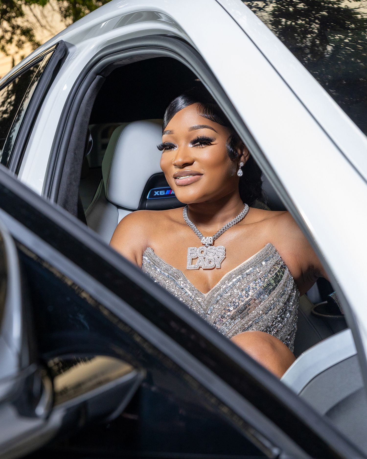 Glamorous woman in silver sequined dress sitting in luxury white SUV, wearing diamond necklace.