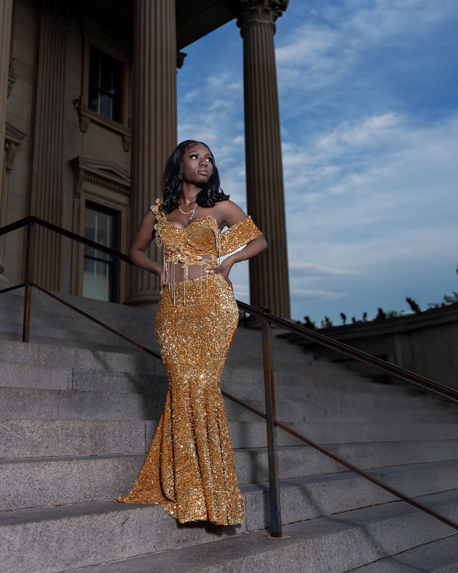 Woman in stunning gold sequin mermaid gown poses on grand staircase with classical columns.