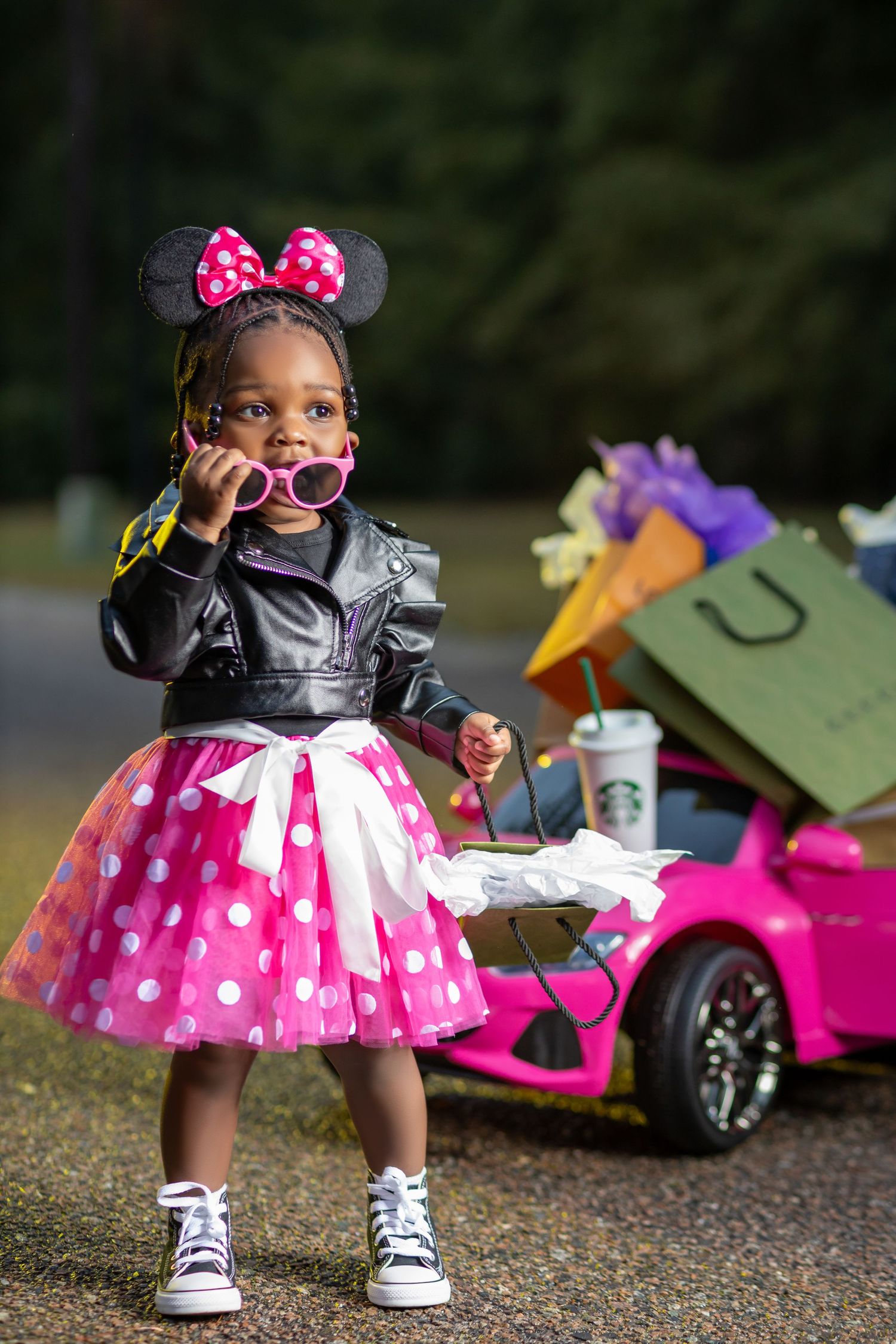 Cute girl in Minnie Mouse costume with sunglasses poses near pink toy car.