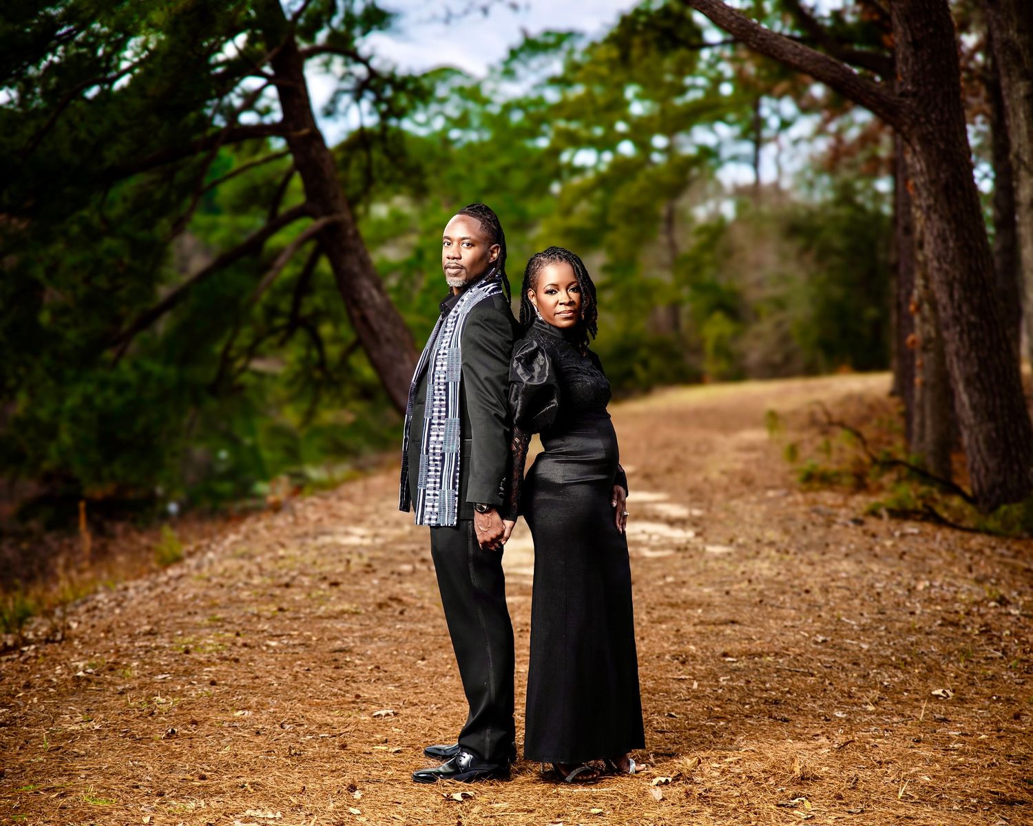 Couple in elegant black attire holding hands back-to-back on a wooded dirt path surrounded by tall trees.