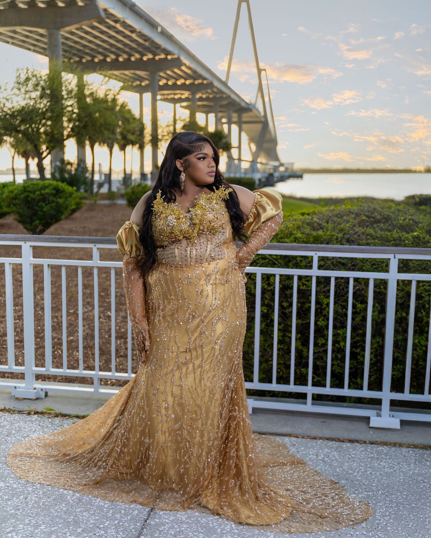 Woman in gold embellished gown posing on waterfront terrace with bridge at sunset.