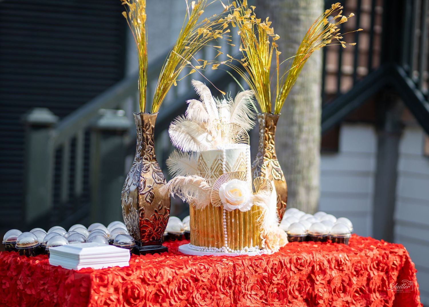 Elegant white and gold celebration cake with feathers displayed on red tablecloth outdoors.