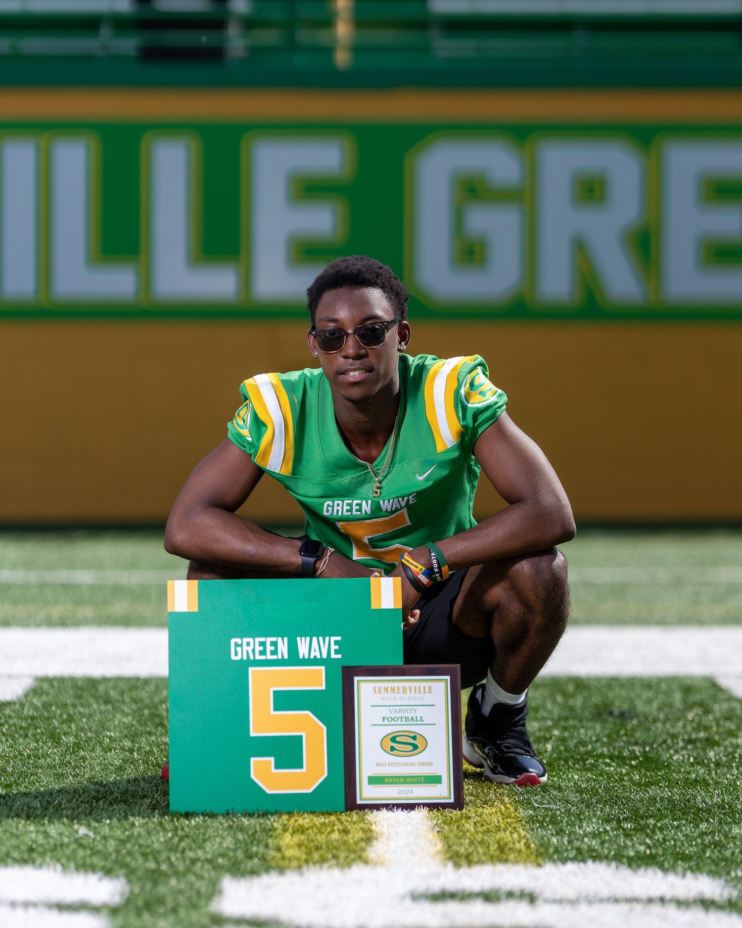 High school football player in a Green Wave jersey posing beside his number 5 sign and football award
