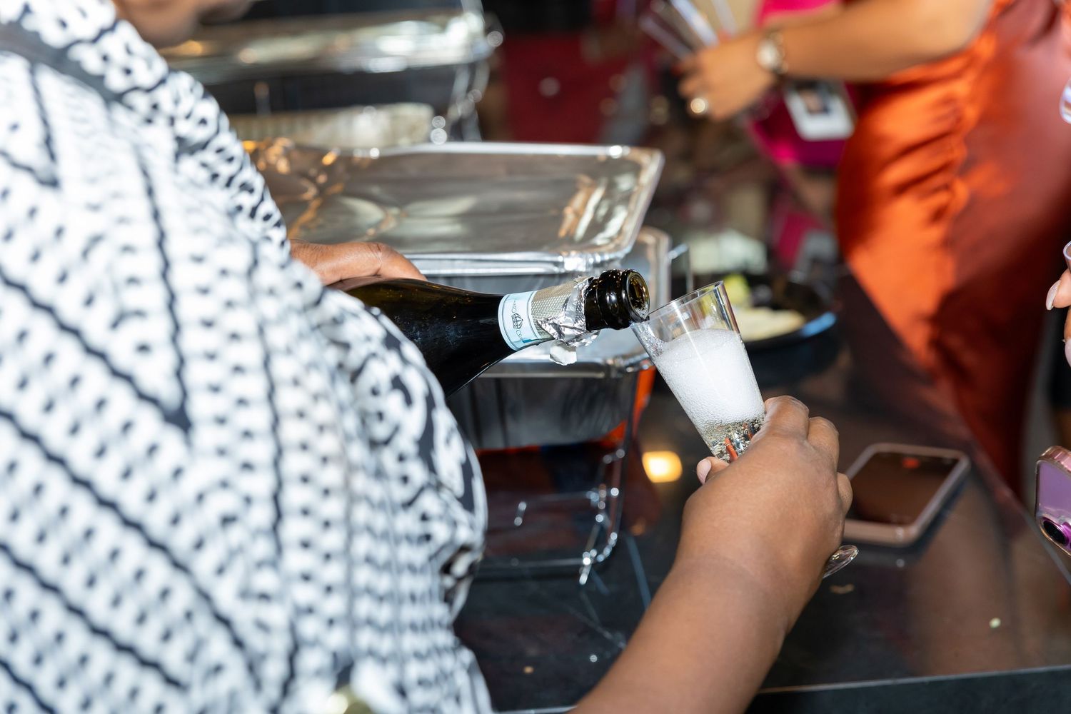 Person in patterned shirt pouring champagne at a party bar setup.