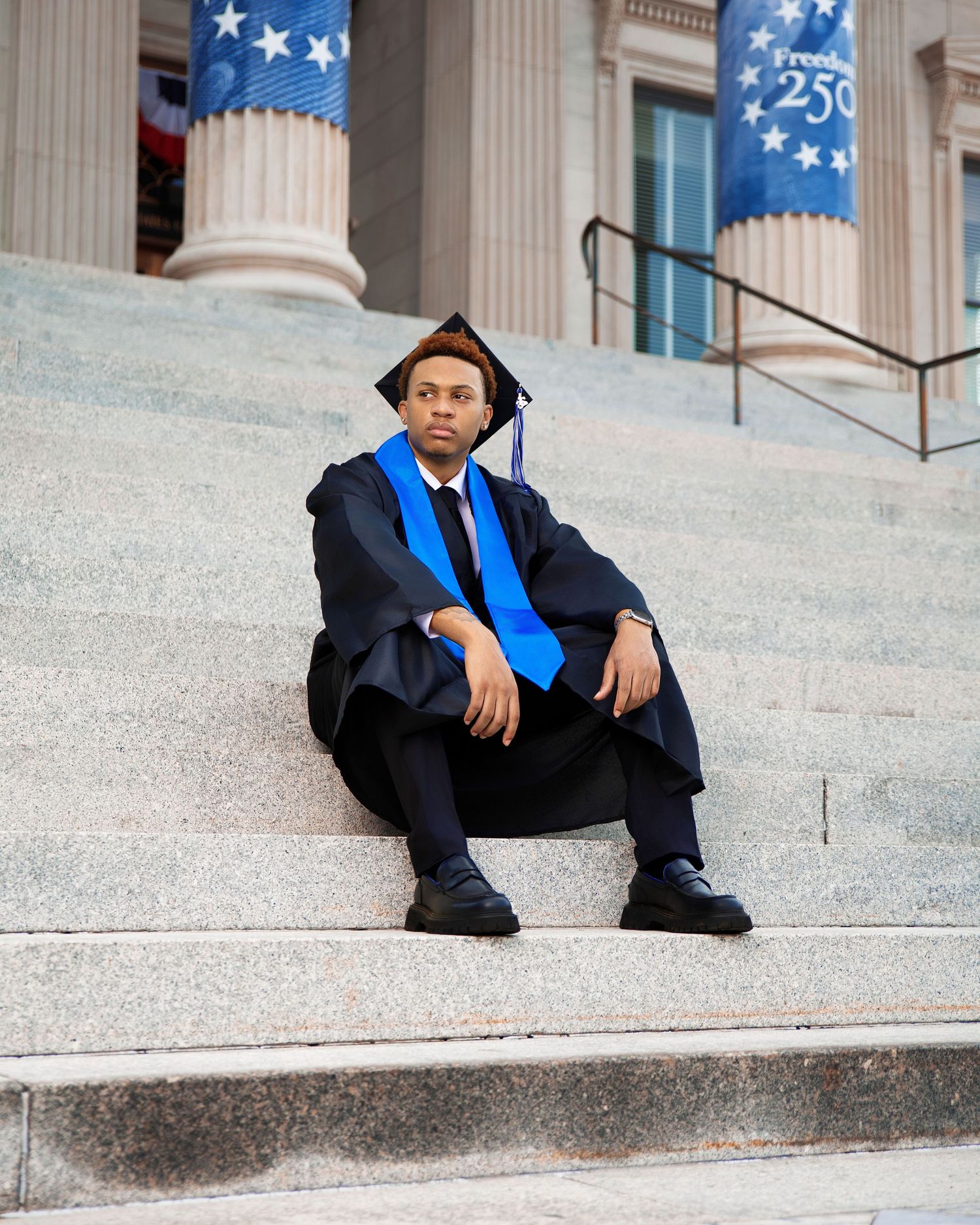 Graduate in black cap and gown sits on steps of a building with blue banners.