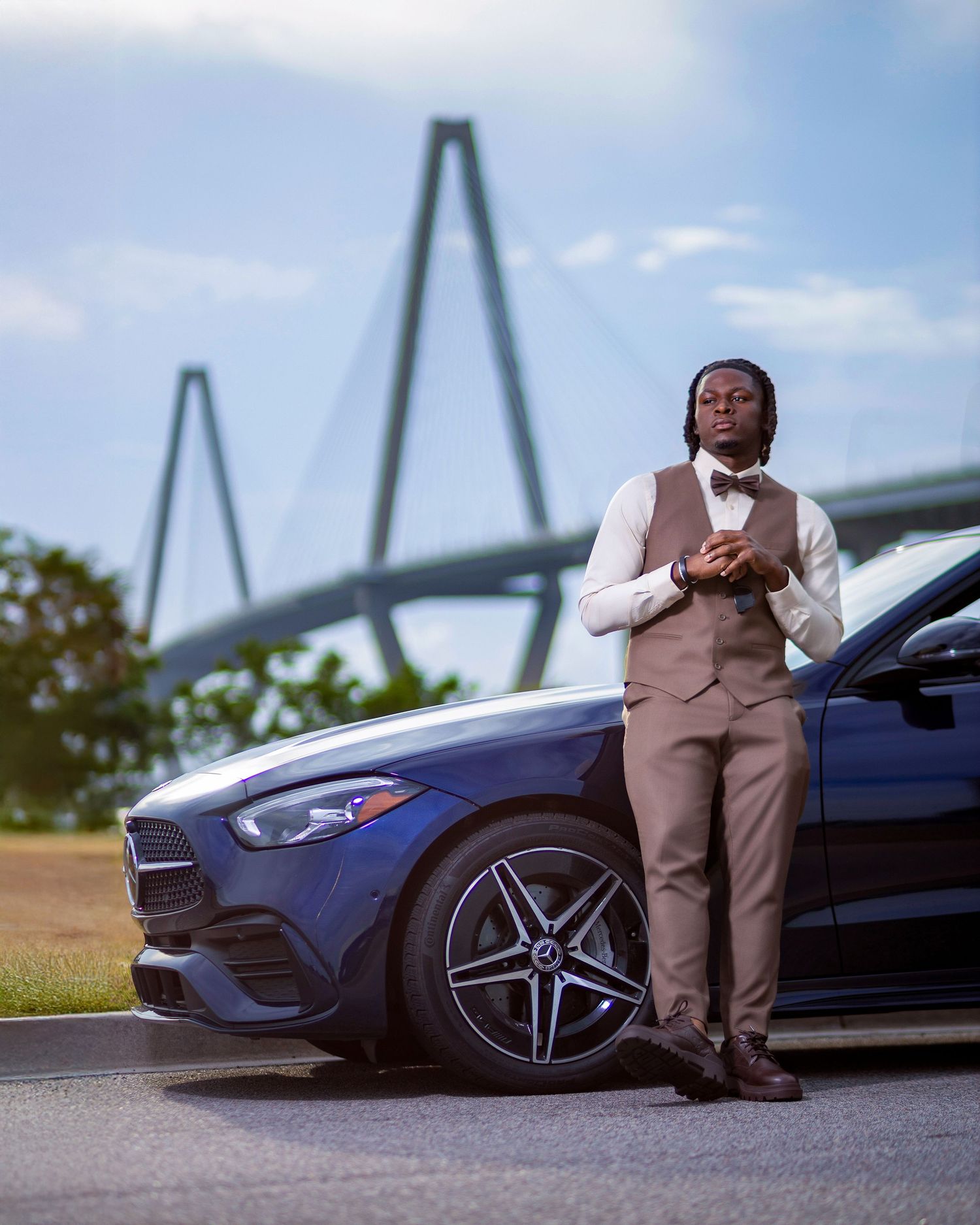 Dapper man in brown vest and bow tie posing beside blue Mercedes with bridge backdrop.